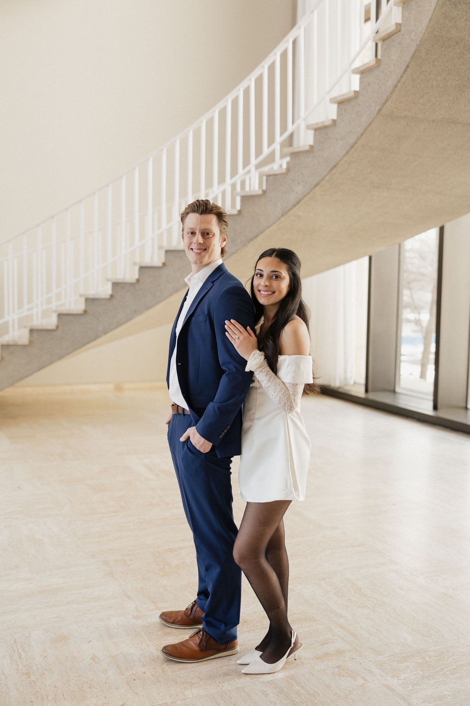 Couple posing playfully near white staircase in elegant indoor setting.
