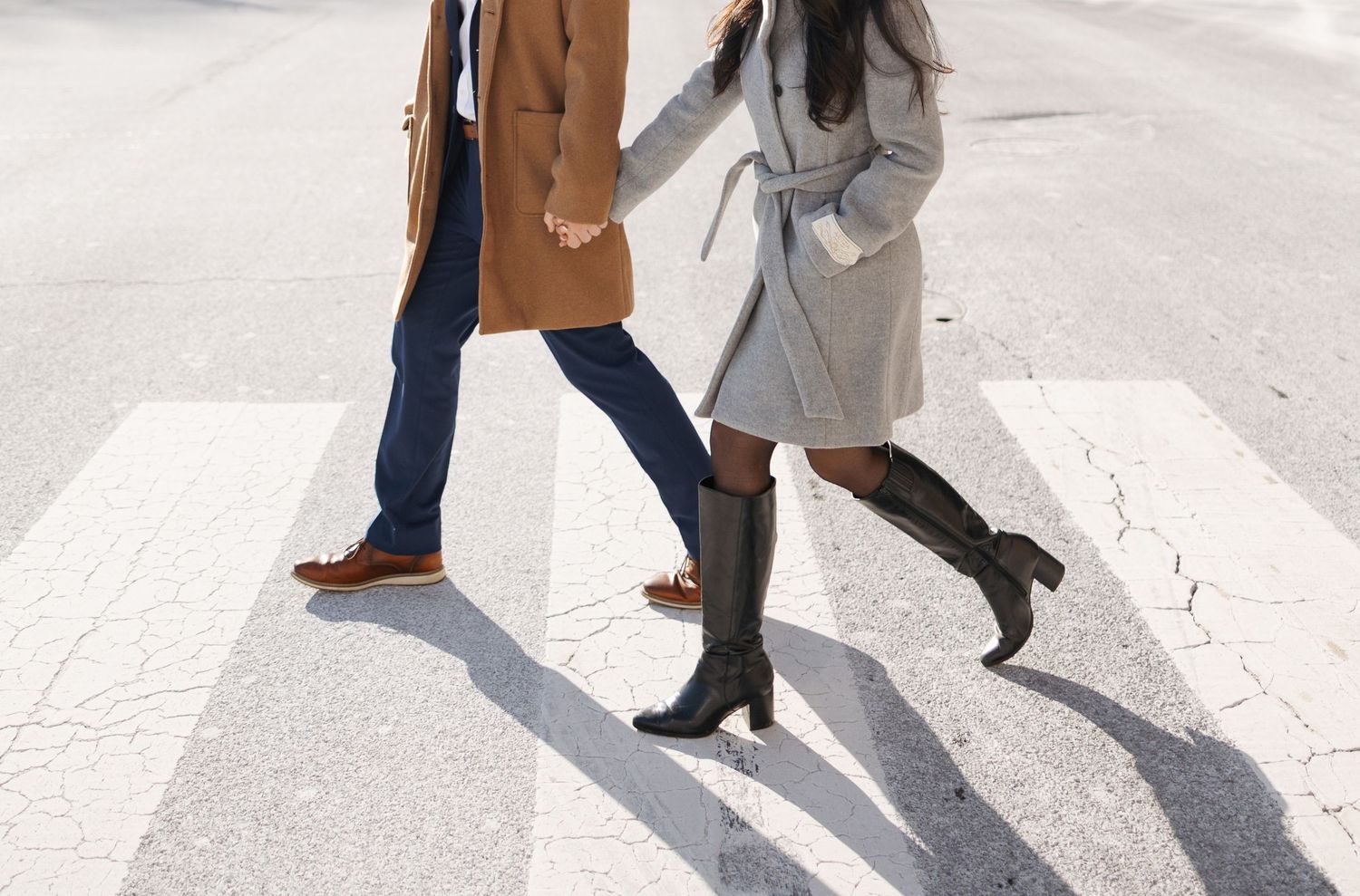 People walking on a white crosswalk casting long shadows on the pavement in bright sunlight.
