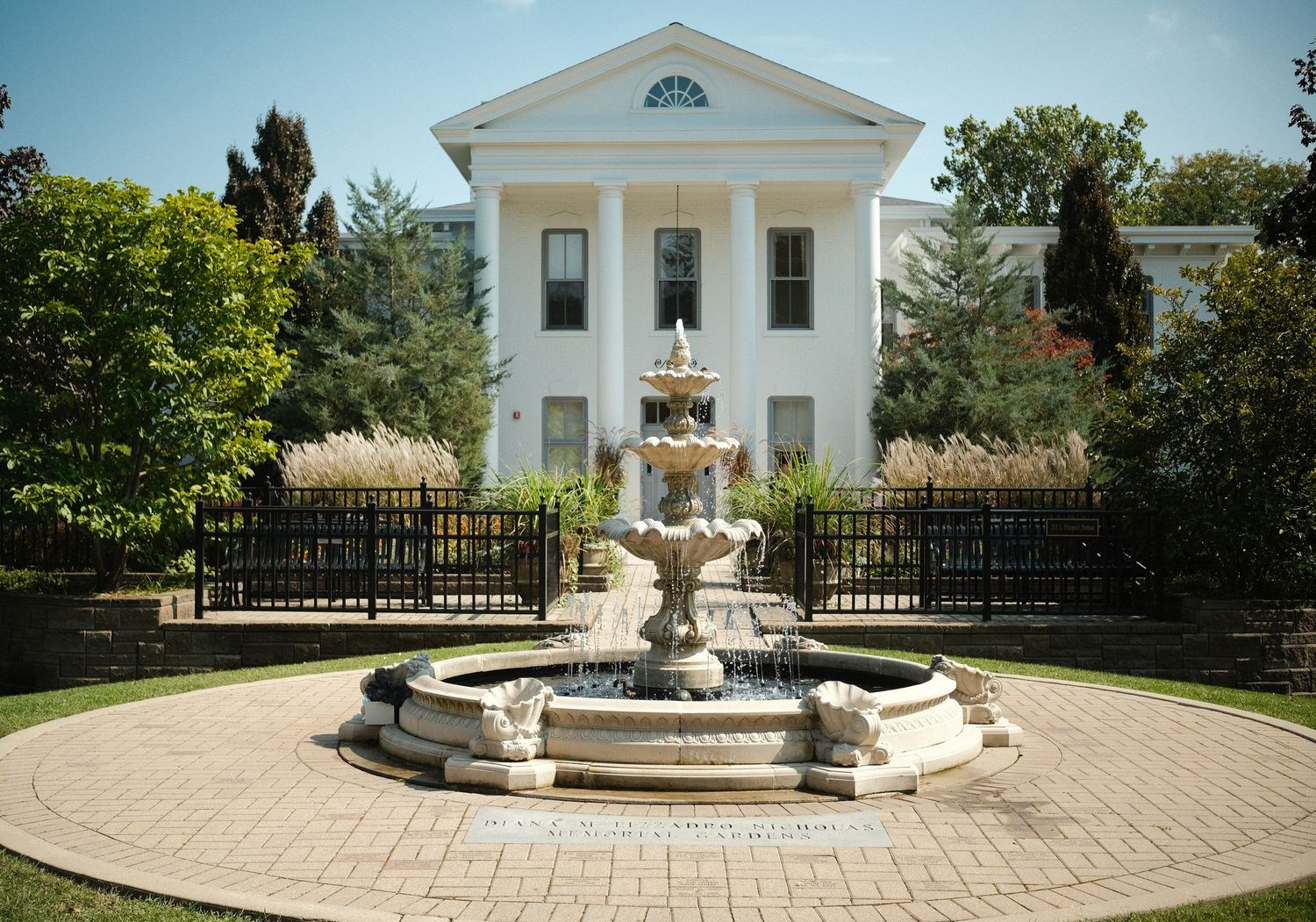 A panoramic view of an elegant white mansion with Greek Revival architecture and a decorative fountain in the landscaped garden.