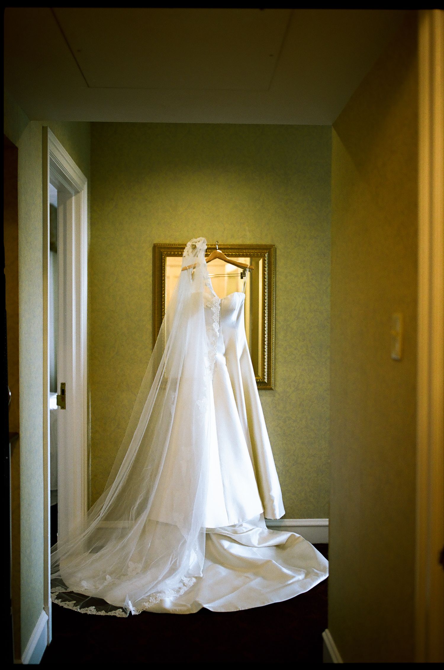 Elegant white wedding dress with long veil hanging on a mirror in a softly lit hotel room.
