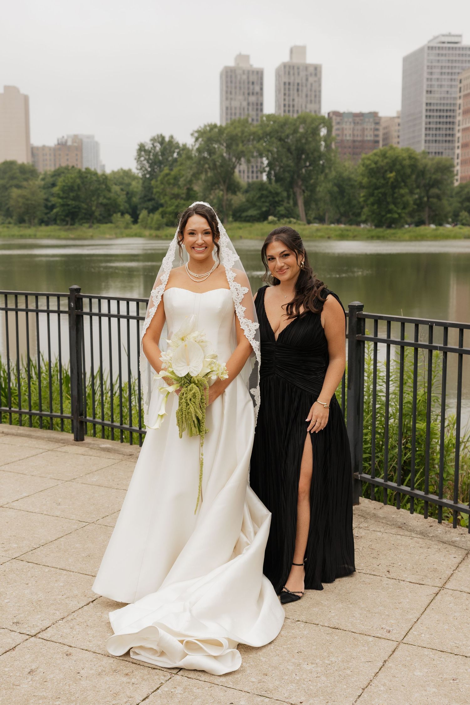 Bride in white gown and veil with bridesmaid in black dress posing by Central Park lake, NYC.
