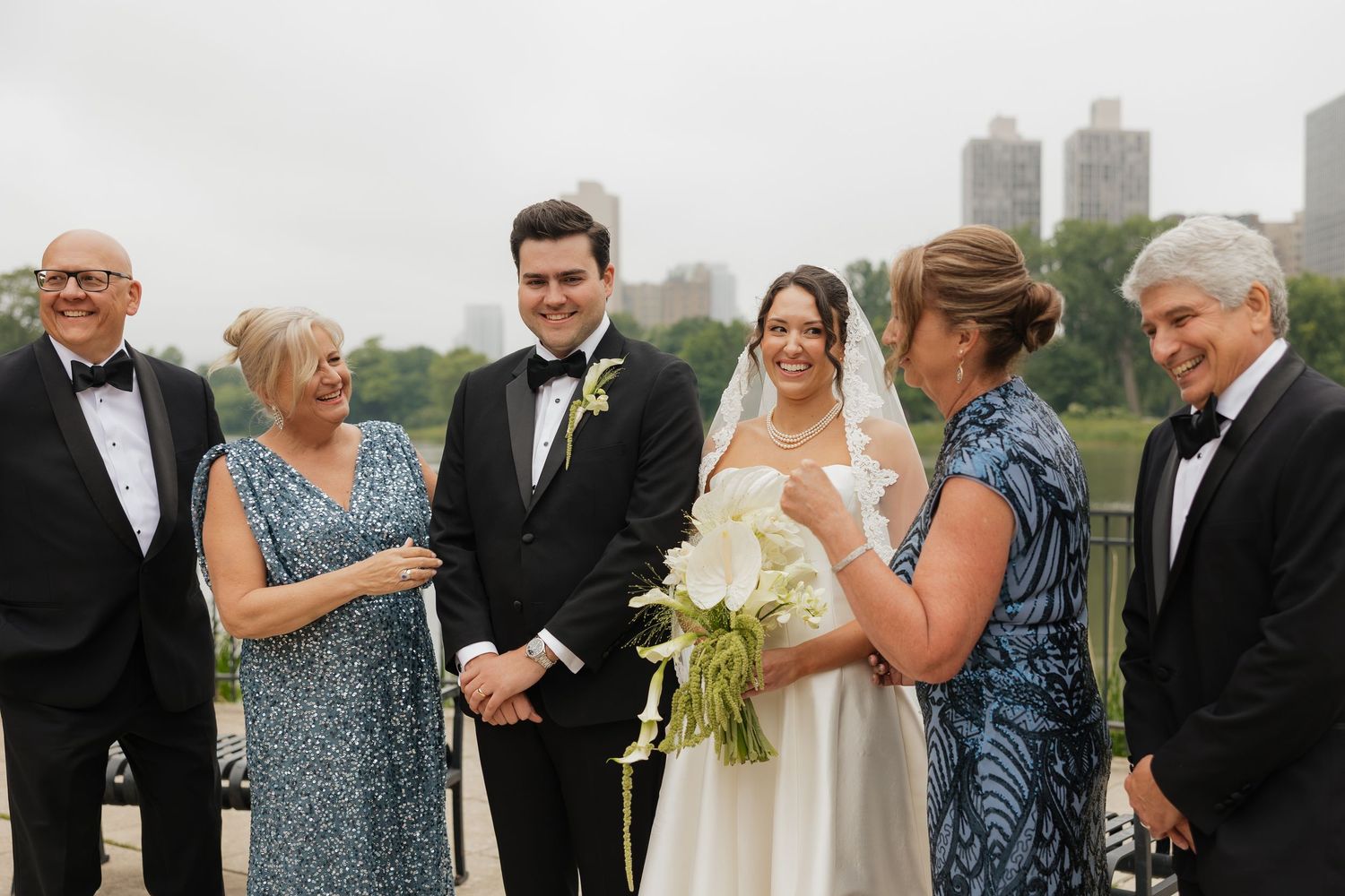 Wedding party smiling outdoors with NYC skyline backdrop, bride holding white bouquet in elegant gown beside groom in tuxedo.