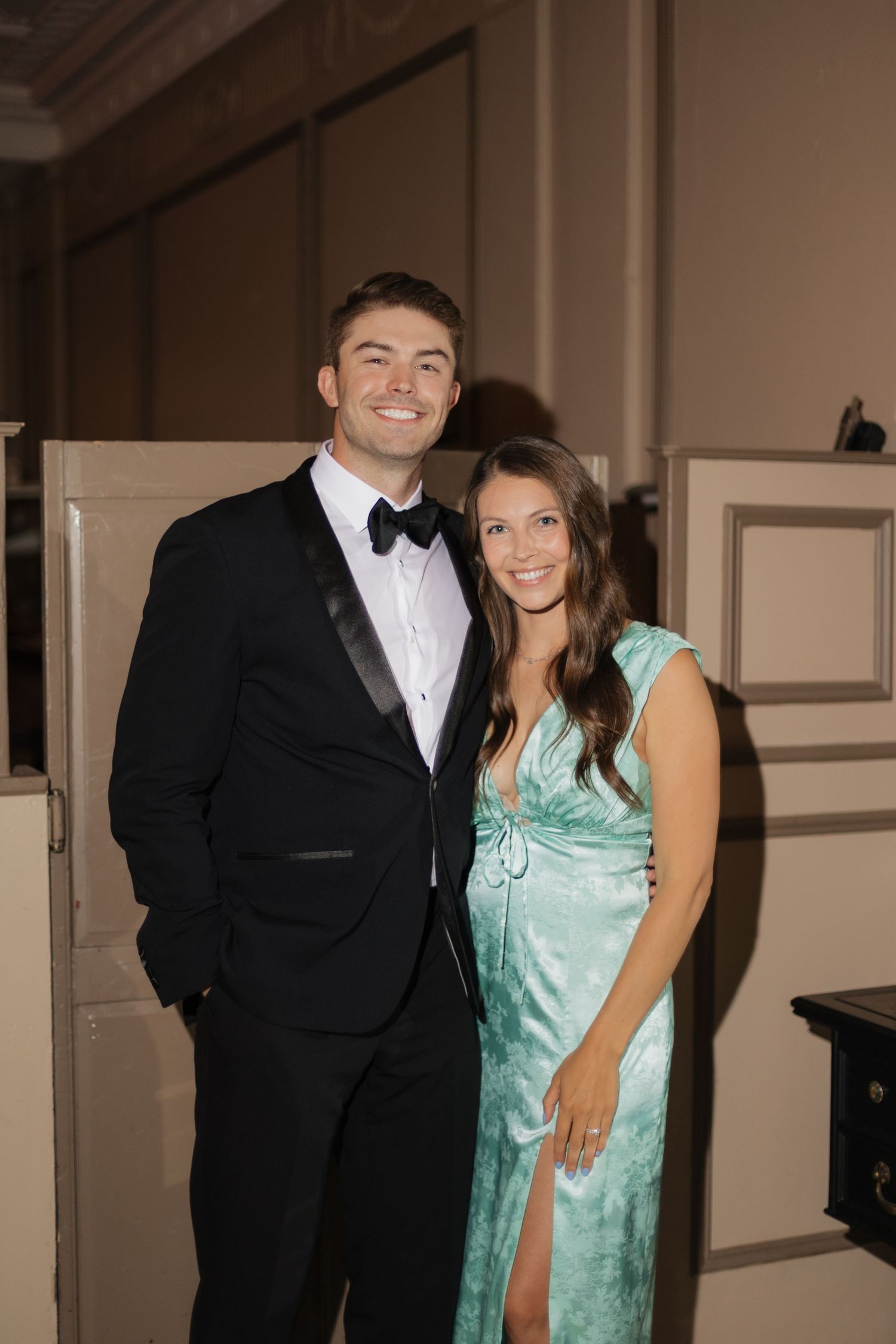 A couple posing at a formal event, man in black tuxedo with bow tie, woman in teal satin gown.