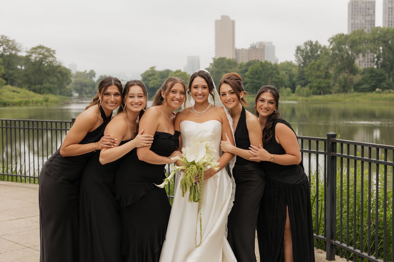 Bride in white gown poses with five bridesmaids in black dresses by a river in a city park.