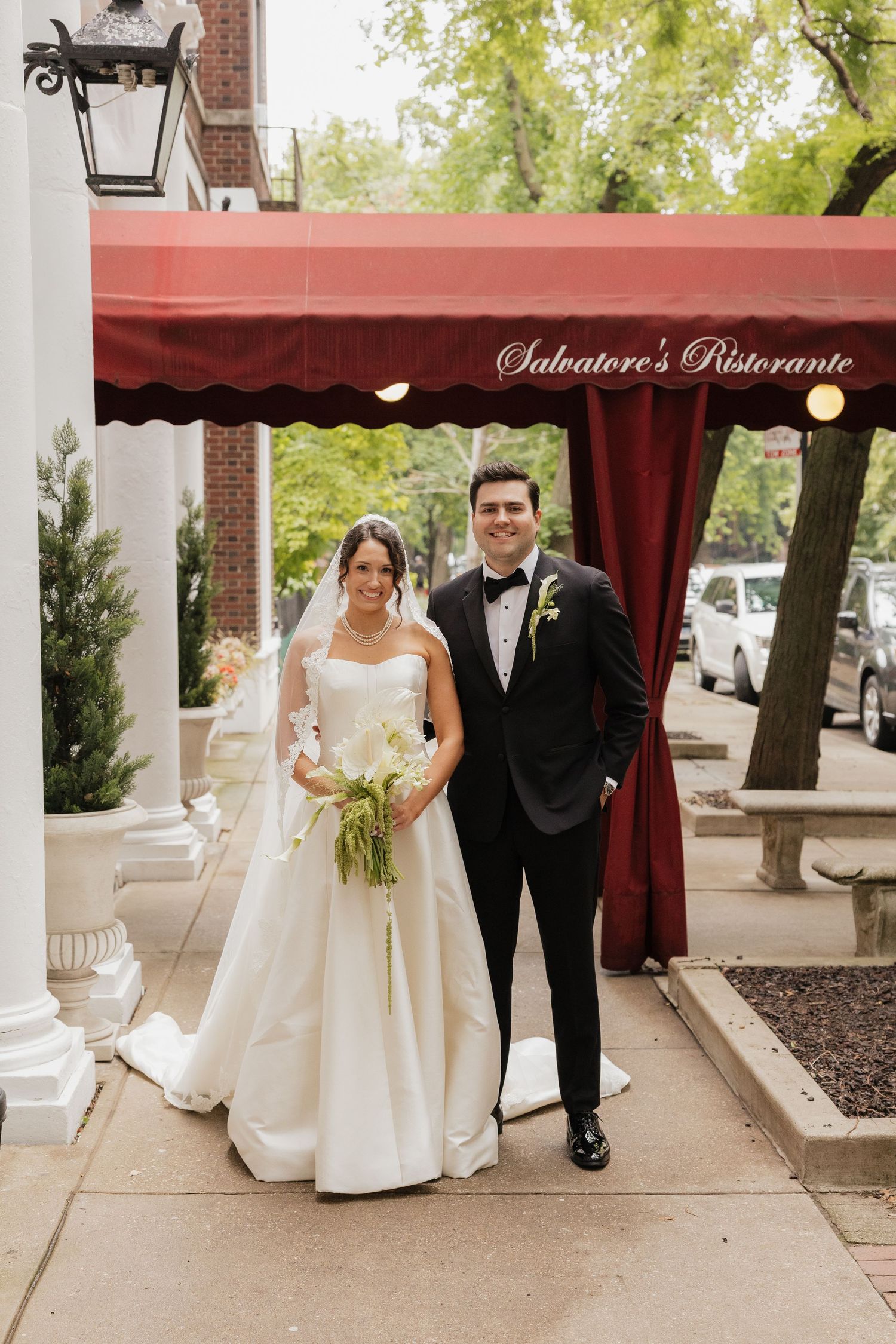 Bride and groom posing outside Salvatore's Ristorante under a red awning, bride holding green bouquet.