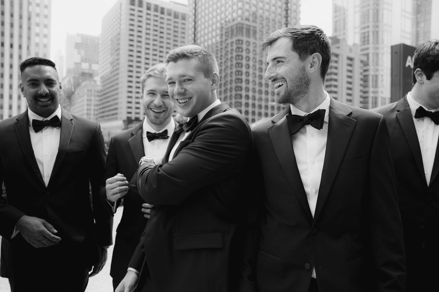 Black and white photo of groomsmen in tuxedos laughing together with city skyscrapers in the background.