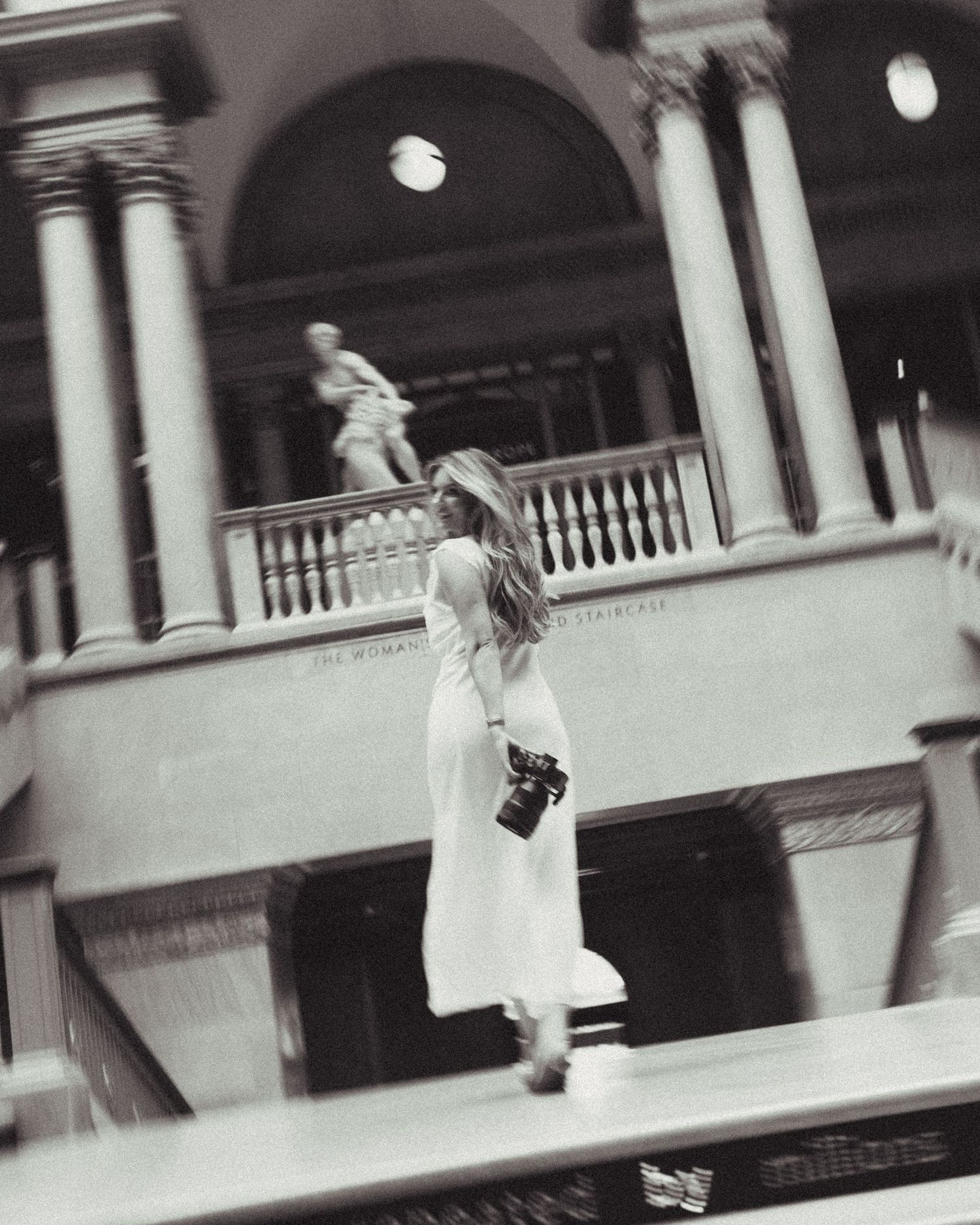 Black and white photo of a woman in a white dress holding a camera inside a grand classical building with columns.