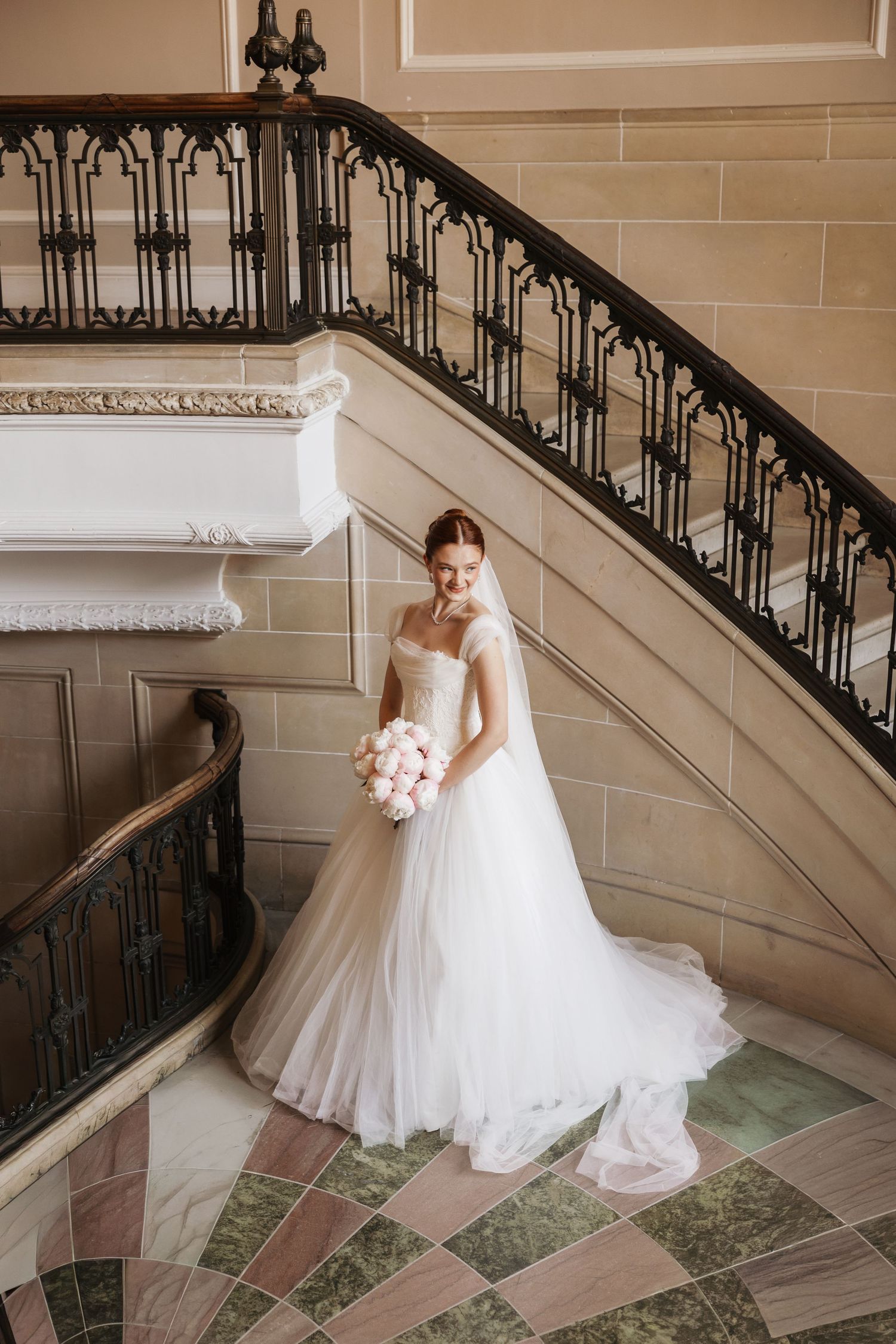 Elegant bride in white off-shoulder gown holding pink bouquet stands on marble floor beside ornate staircase.