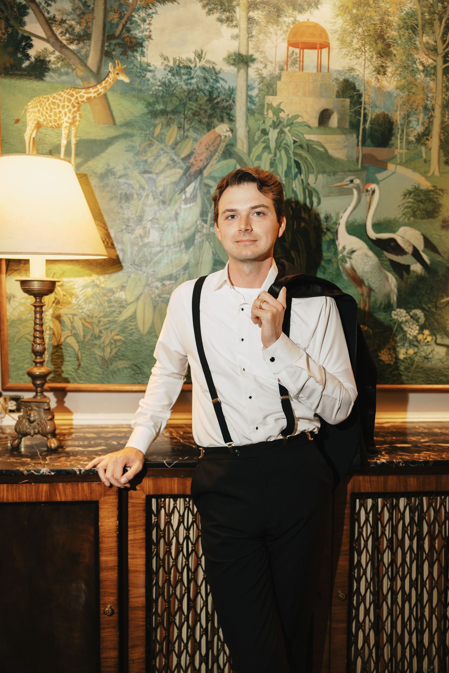 Elegant man in white shirt with suspenders holding suit jacket in ornate room with tropical mural and giraffe artwork.