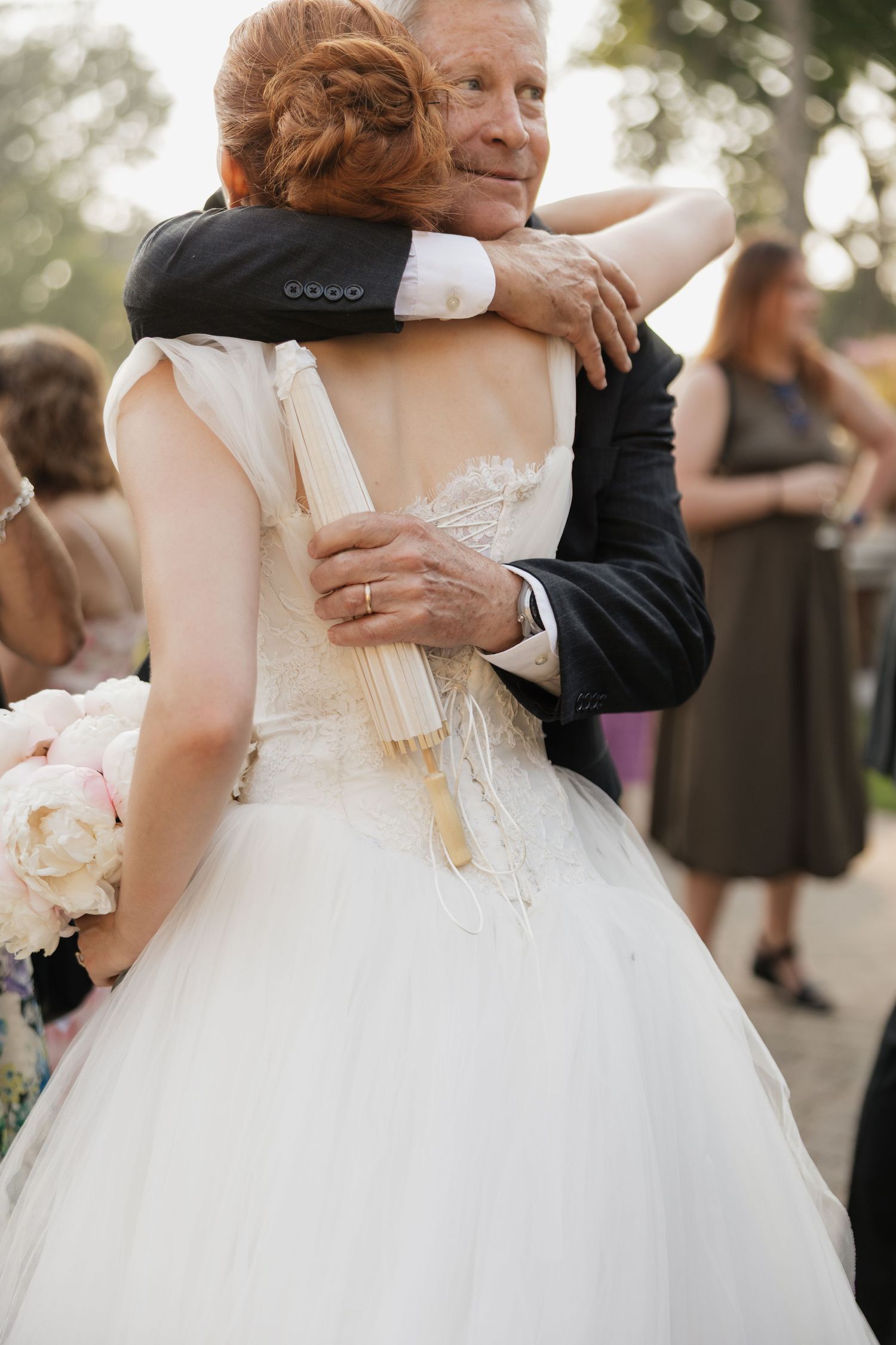 Bride in white lace gown embracing an older man in a suit at an outdoor wedding reception.