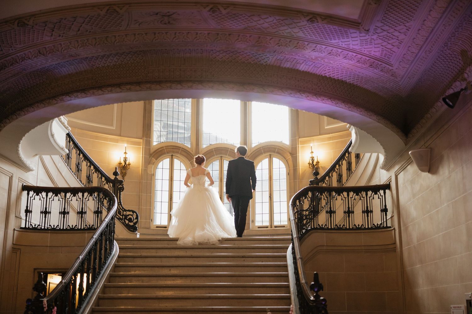 Bride and groom stand atop grand staircase in elegant venue, arched ceiling and ornate railings framing romantic wedding portrait.