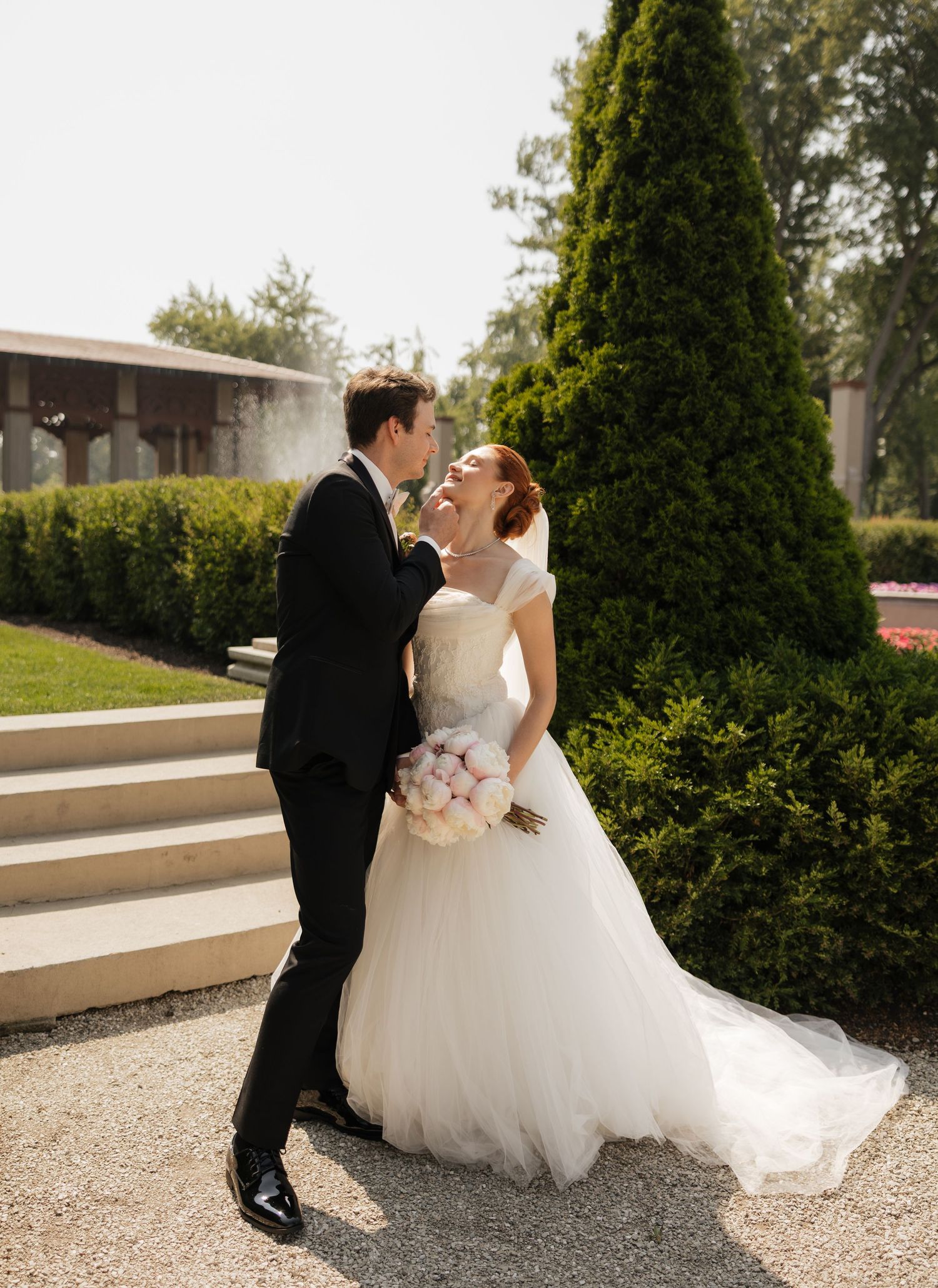 Bride in white ball gown holding pink bouquet laughs with groom in black suit in a garden setting.