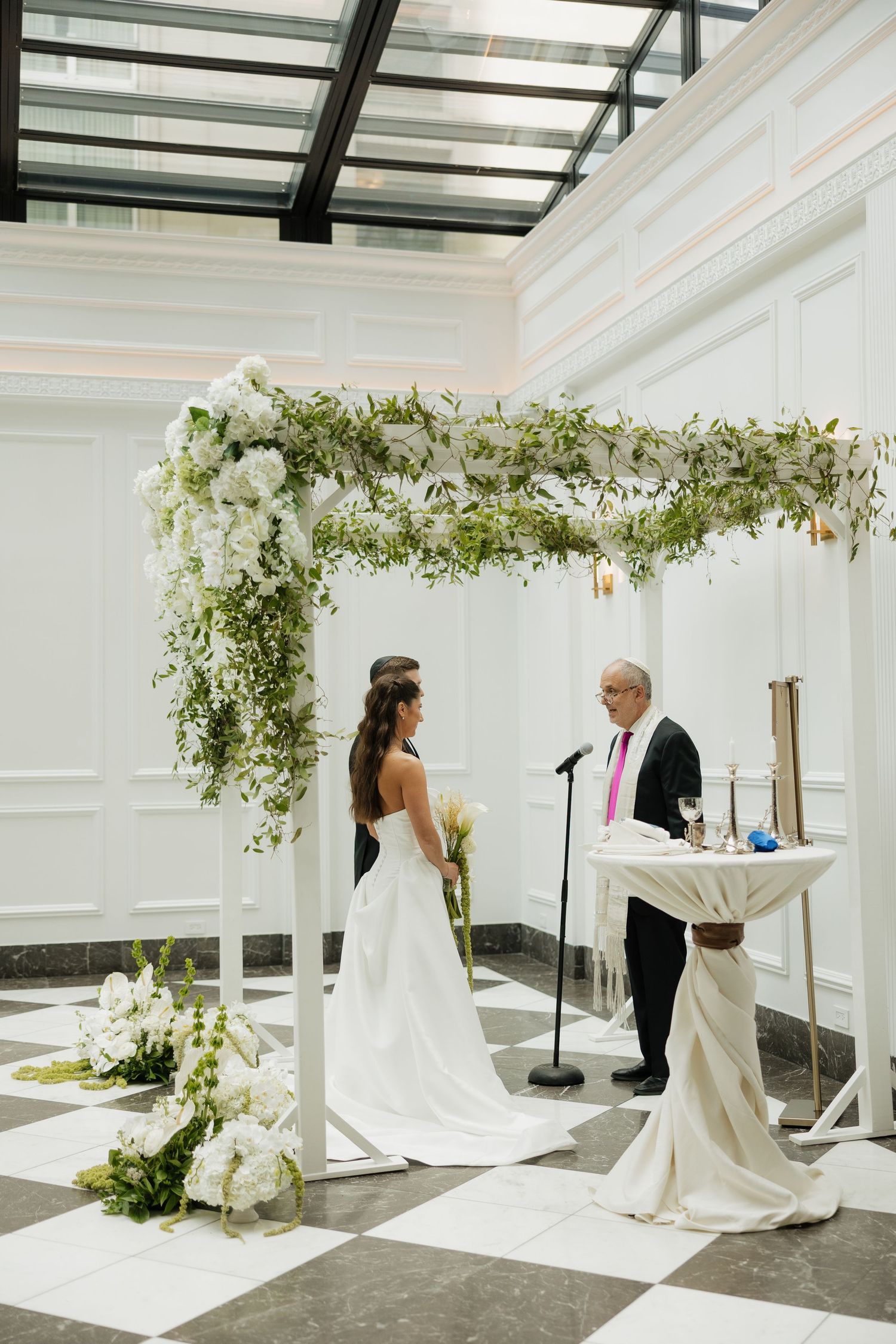 Bride stands under floral chuppah adorned with white flowers and greenery during wedding ceremony.