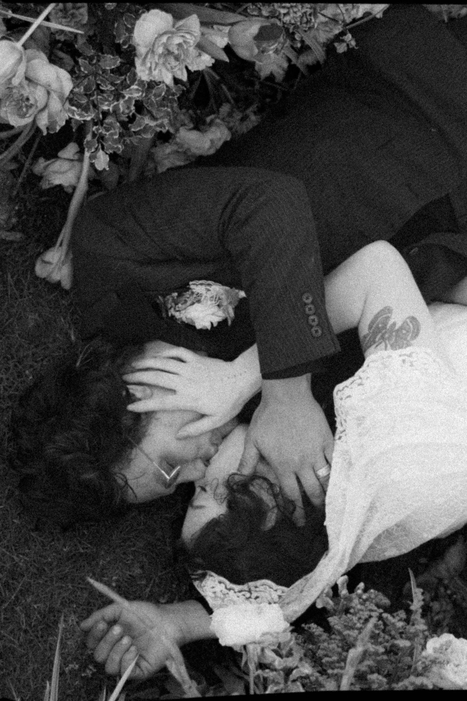 Black and white photo of a couple lying in grass surrounded by flowers, sharing a romantic kiss.