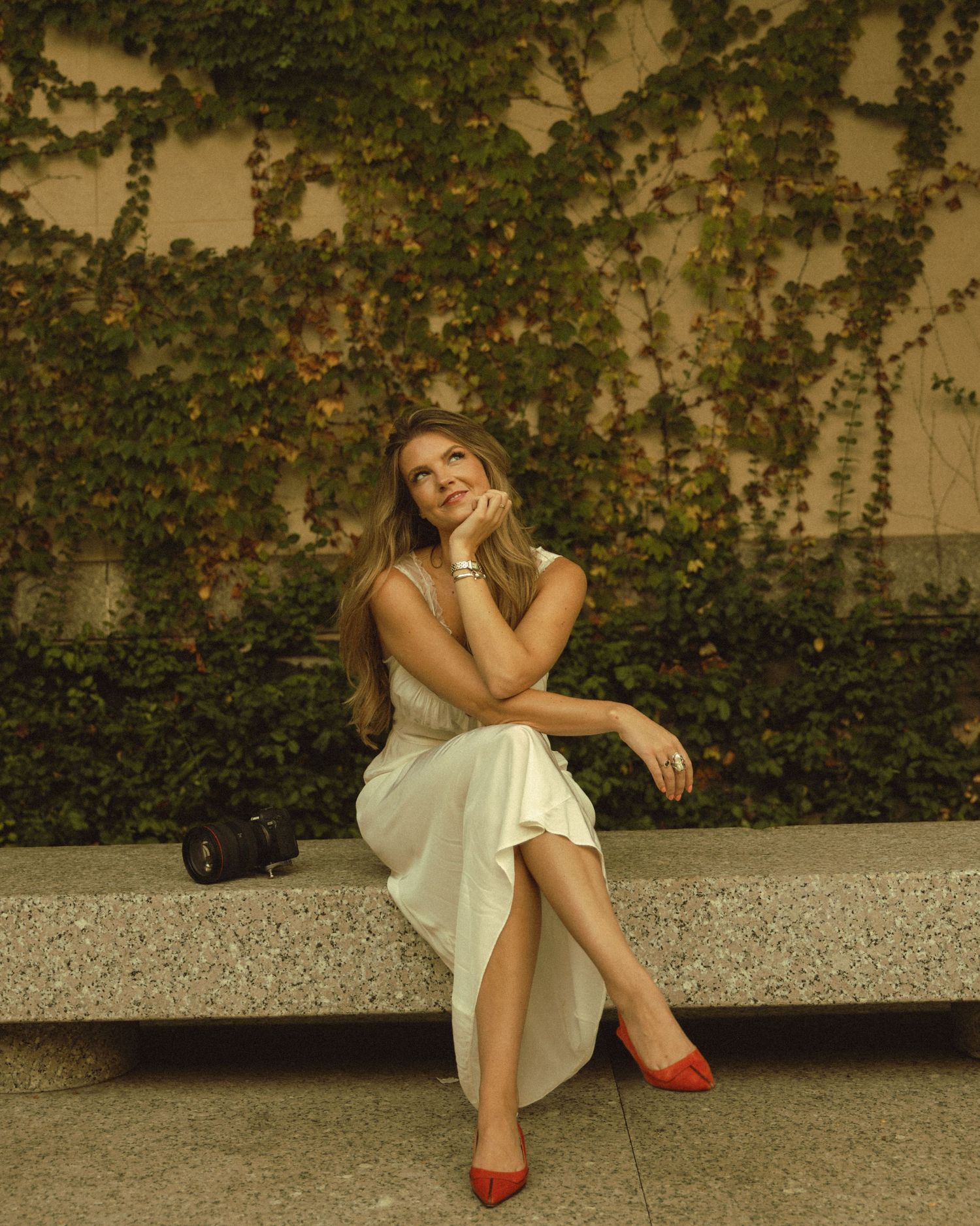 Woman in white dress and red heels sits on stone bench smiling, ivy-covered wall behind her.