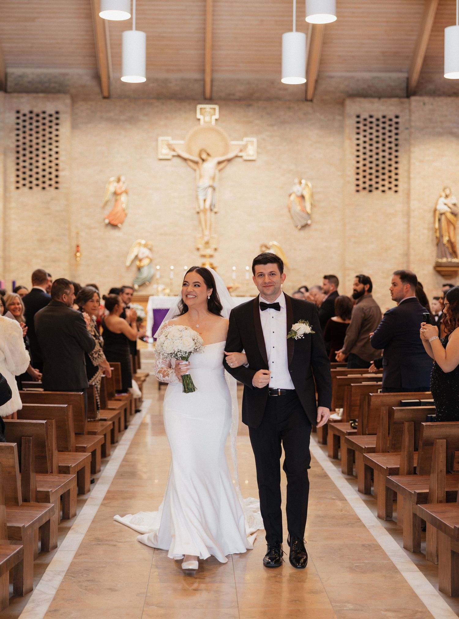 Wedding party exits Catholic church ceremony as guests line wooden pews in modern sanctuary interior.