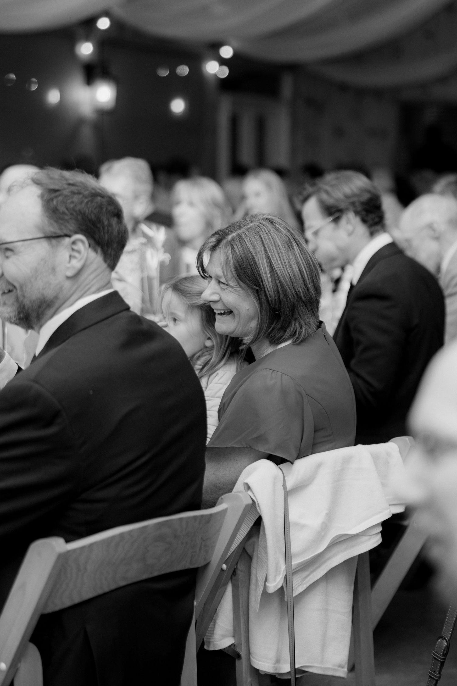 Black and white photo of smiling wedding guests seated in rows under string lights, laughing together during a reception speech.
