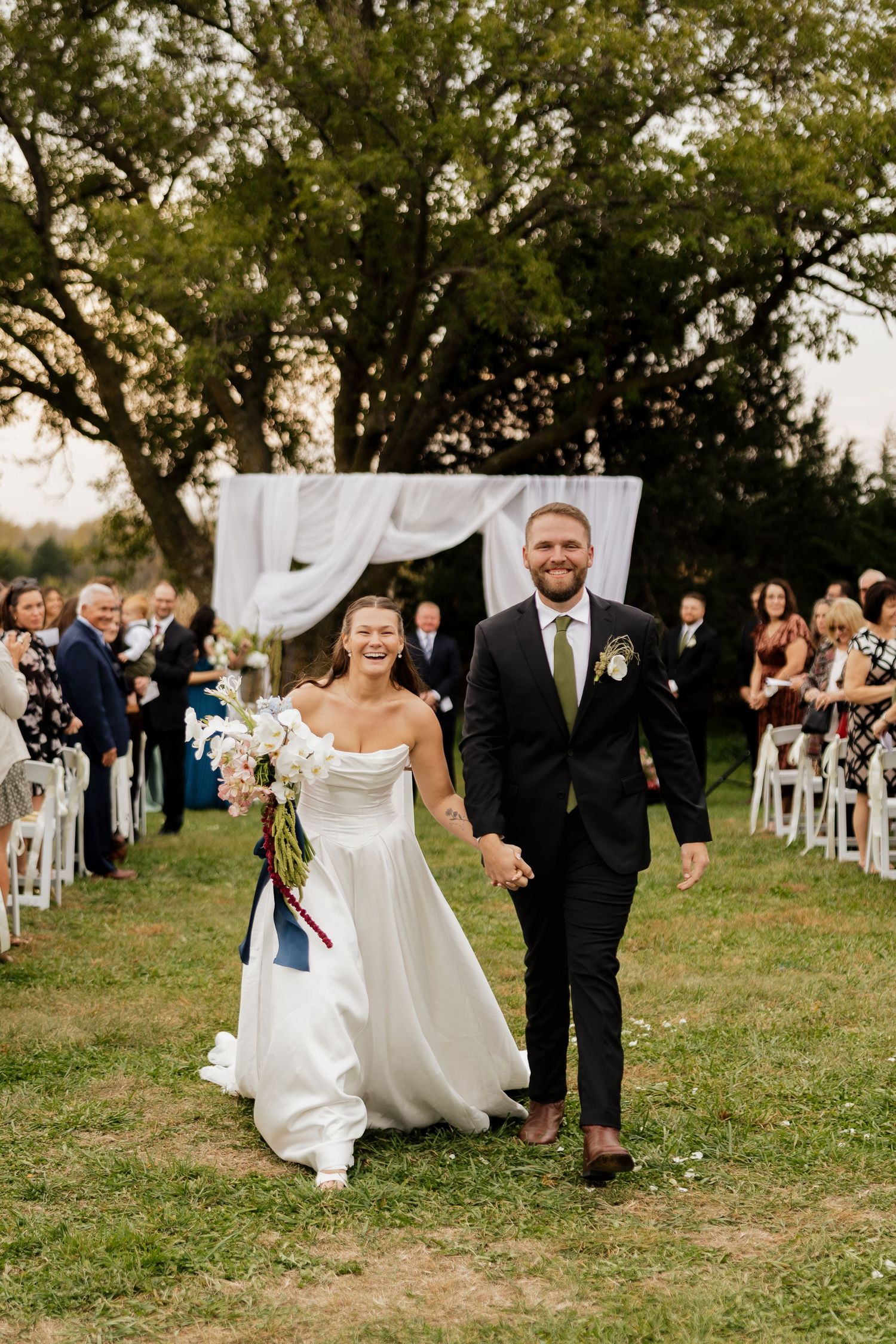 Smiling bride in a white gown and groom in a dark suit hold hands walking down the grass aisle after their outdoor wedding ceremony.
