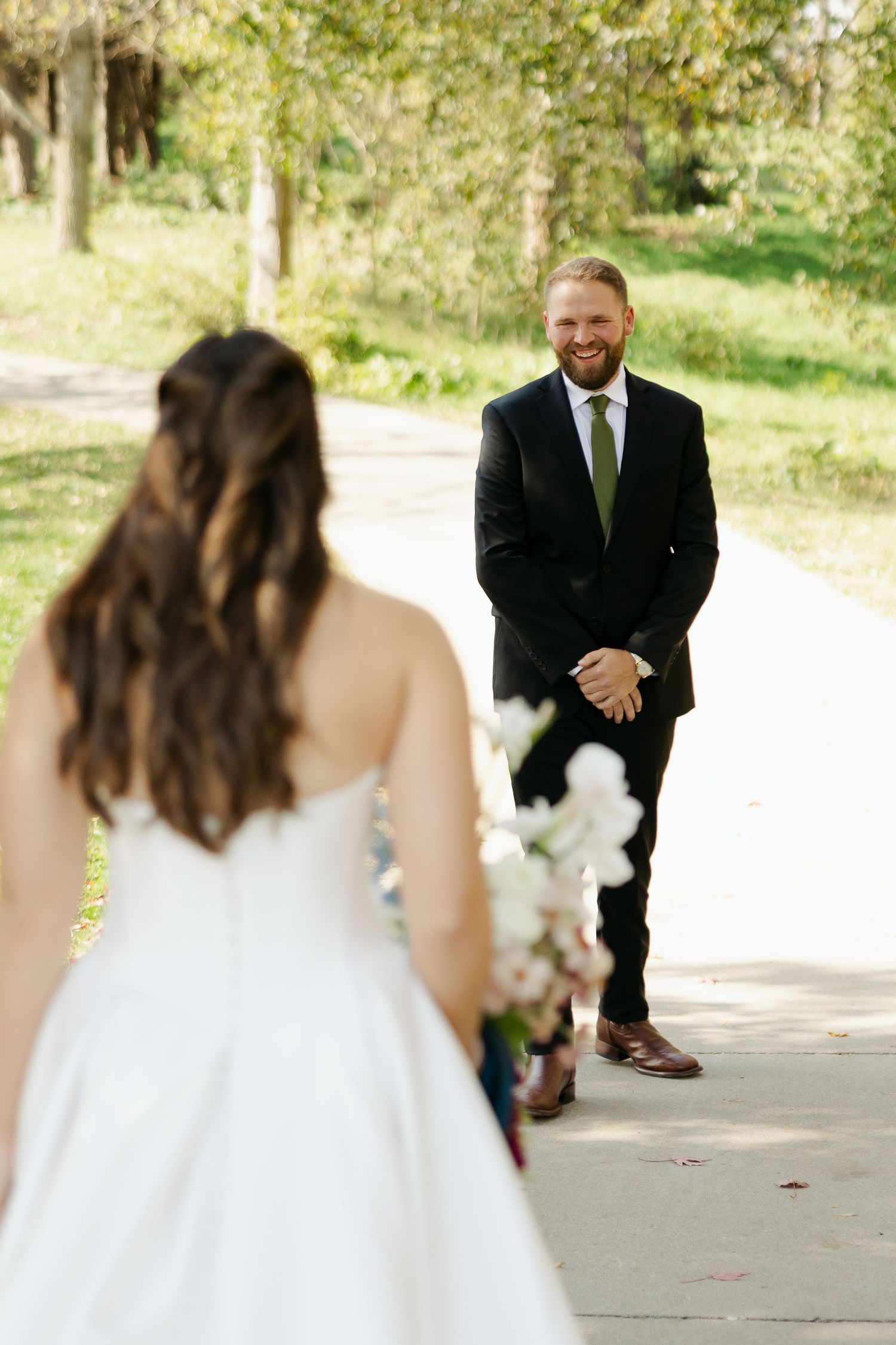 Smiling groom in a black suit and green tie awaits his bride in a white gown holding a bouquet during a first look on a park path.