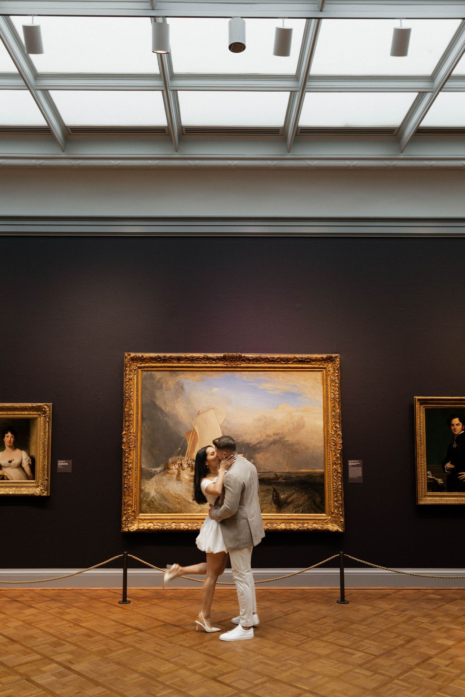 Couple sharing a romantic kiss in an art museum gallery with classic paintings in ornate gold frames.