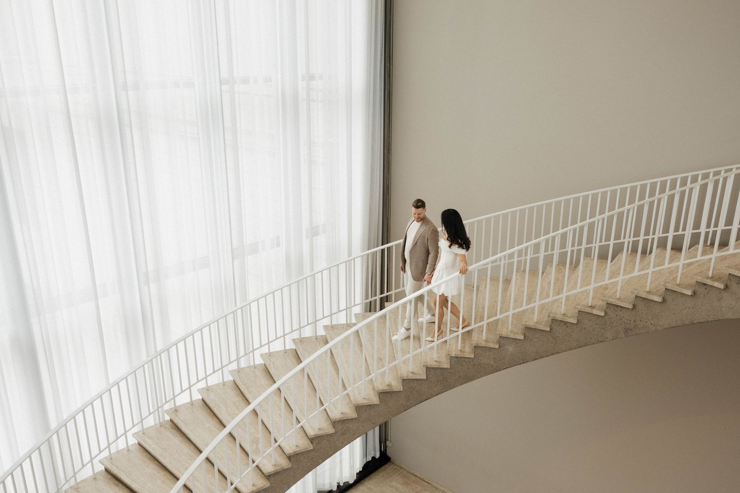 Couple strolling up a curved white staircase in a bright minimalist space with floor-to-ceiling sheer drapes.