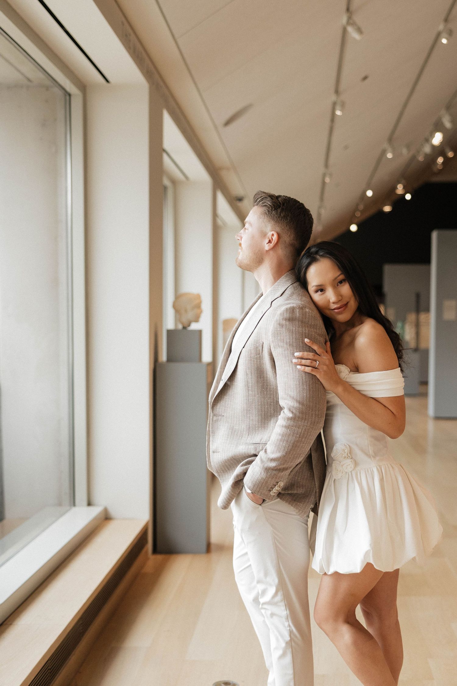 Couple embracing in a museum gallery, man in beige suit, woman in white mini dress.
