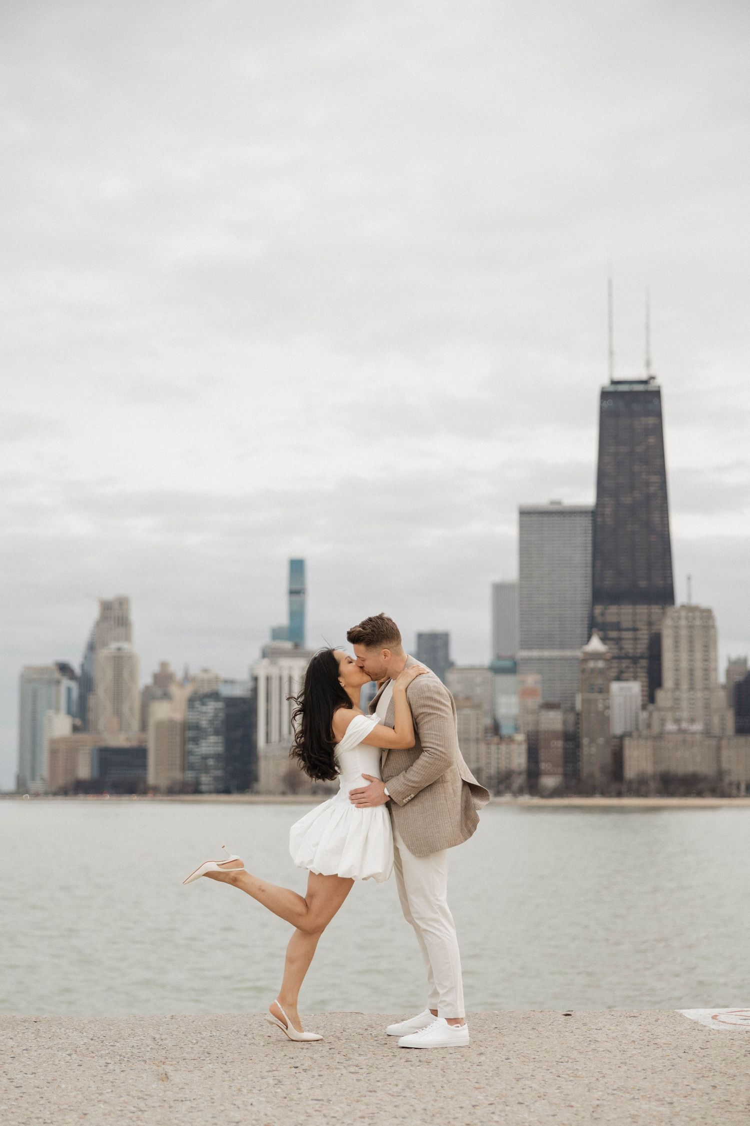 Couple sharing a romantic kiss on Chicago lakefront with city skyline backdrop on a cloudy day.