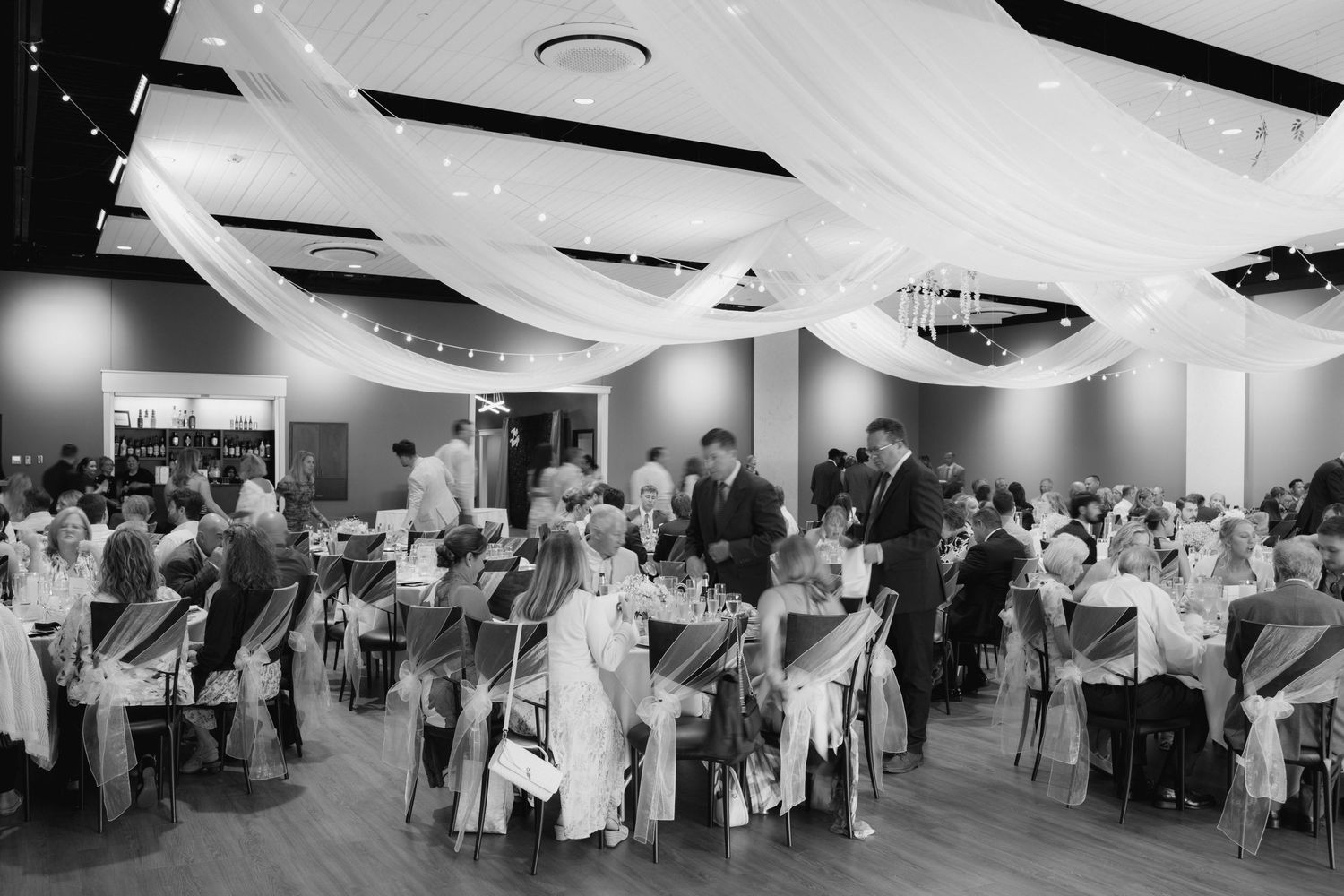 Black and white photo of elegant wedding reception with draped ceiling fabric, round tables, and guests mingling in a modern venue.