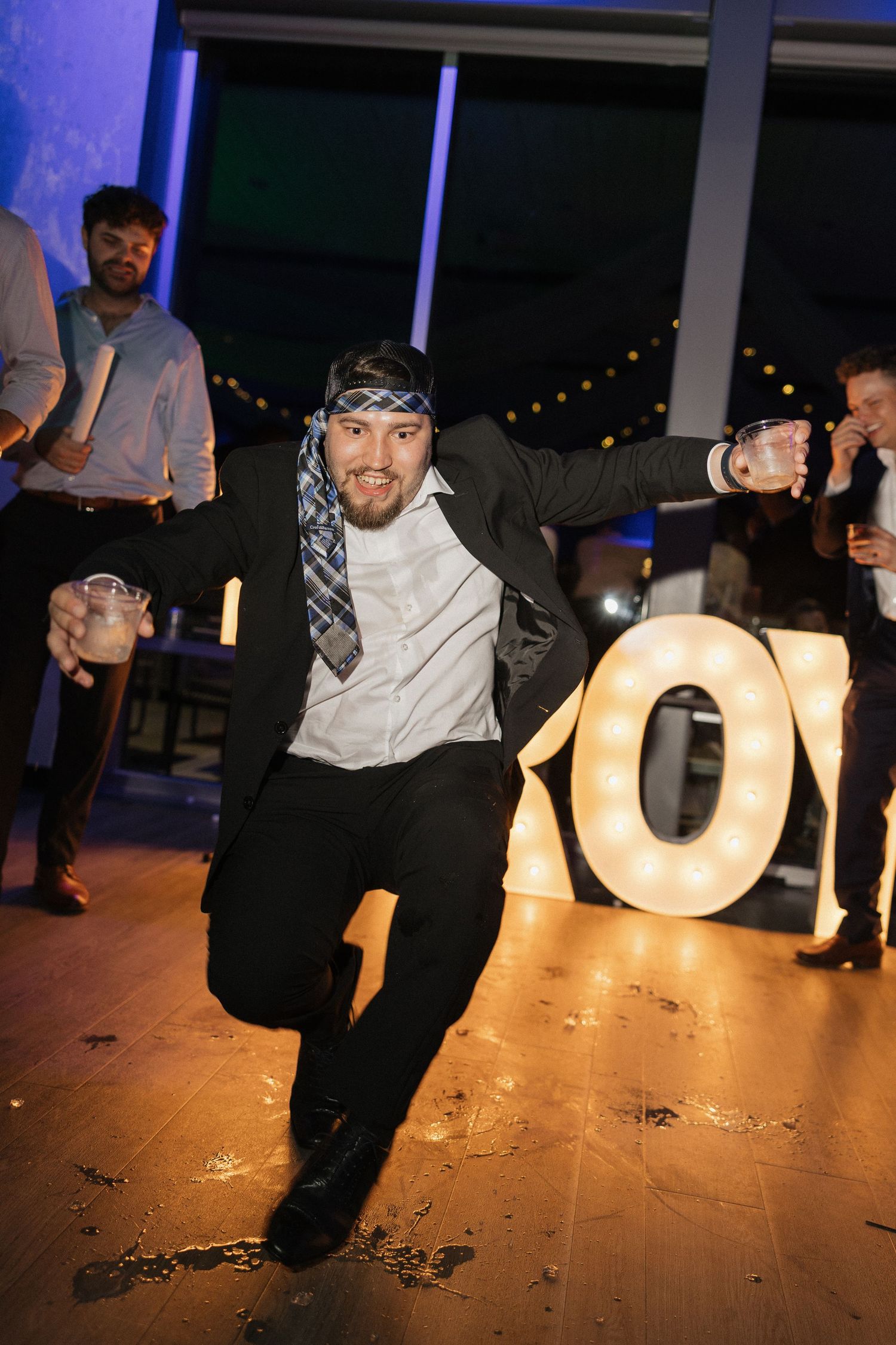 Man dancing energetically at a party near illuminated letter signs, wearing a tie as headband and holding drinks.
