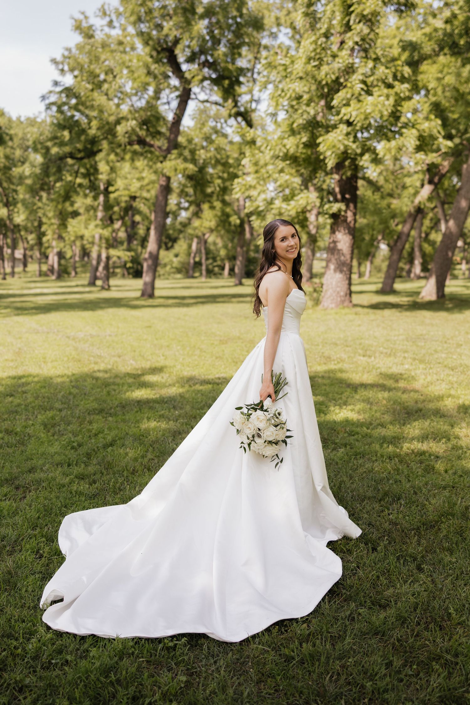 Smiling bride in elegant white ball gown with train holds bouquet in lush green park setting.