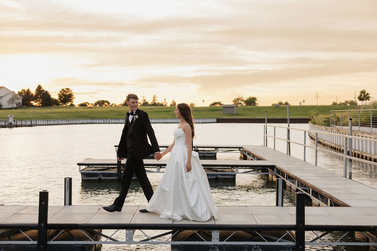 Bride and groom walk hand-in-hand on a waterfront dock at sunset during their wedding day.