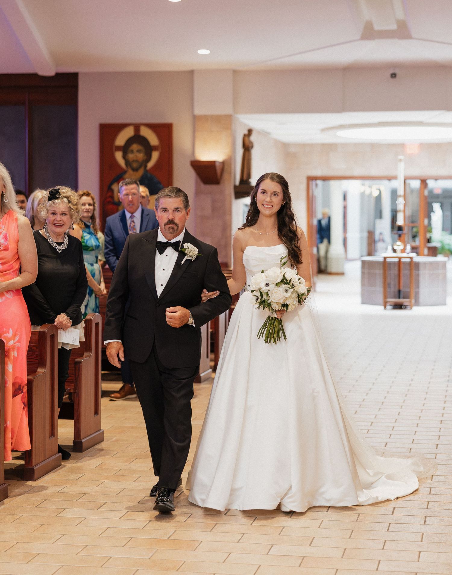 Bride in white ball gown walks down church aisle with escort during elegant wedding ceremony.