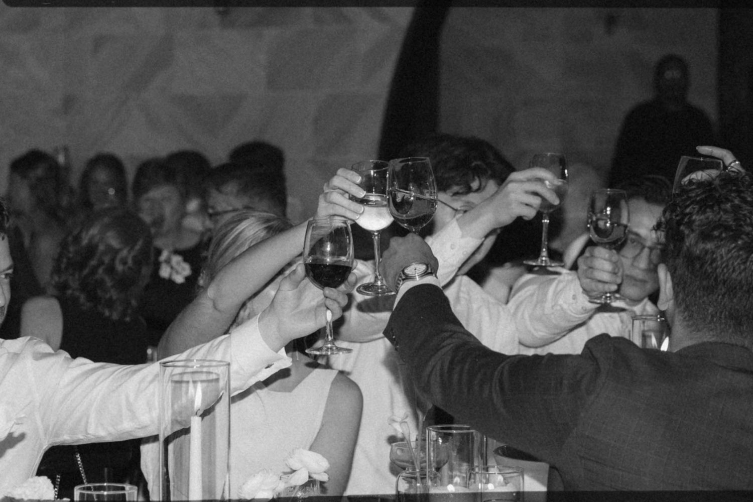 Black and white photo of wedding guests raising glasses in a celebratory toast at a reception dinner.