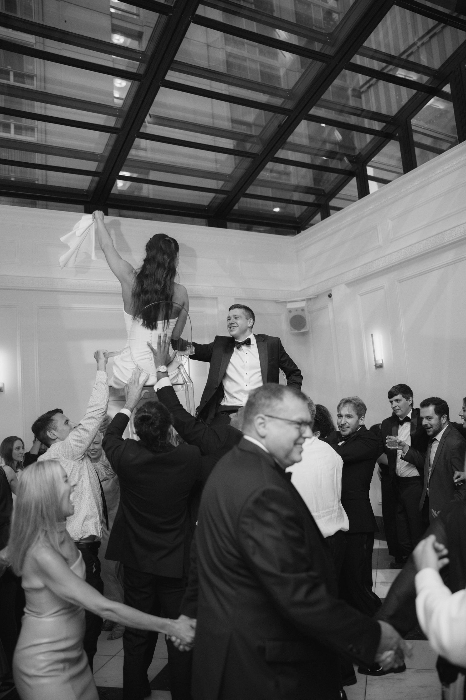 Black and white photo of bride and groom being lifted by cheering guests during traditional hora wedding dance.