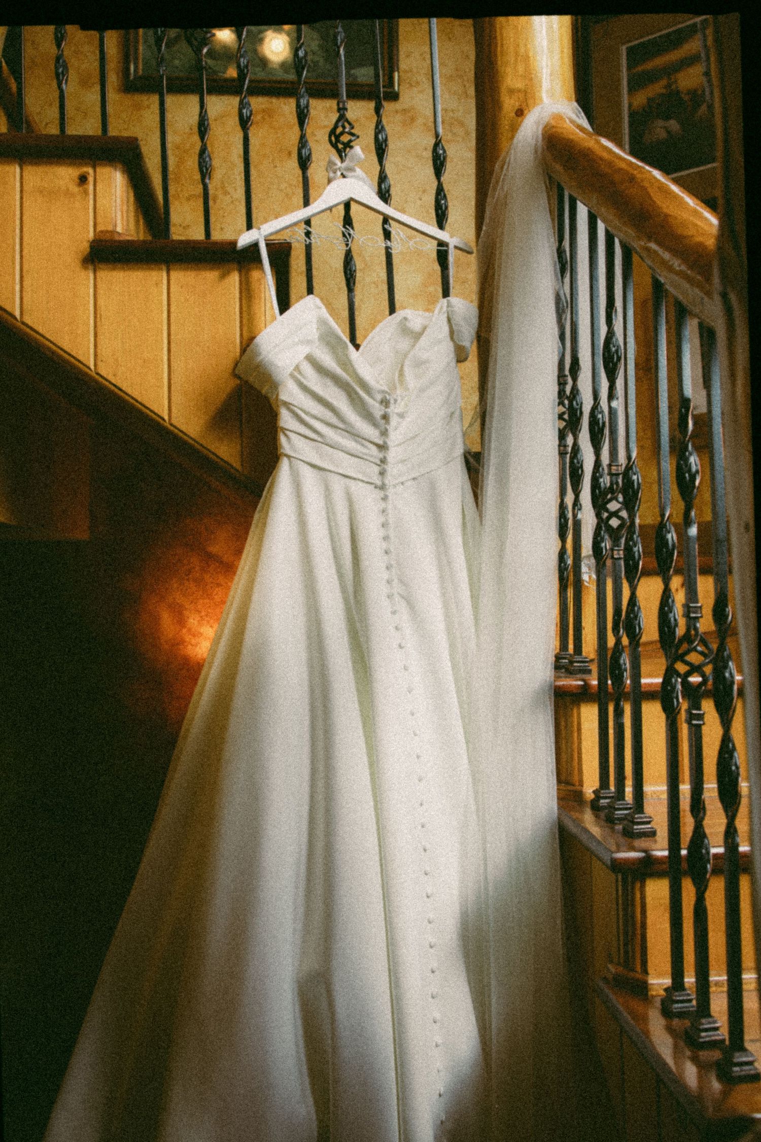Elegant white strapless wedding gown with button detail hanging on a hanger over a wooden staircase railing.