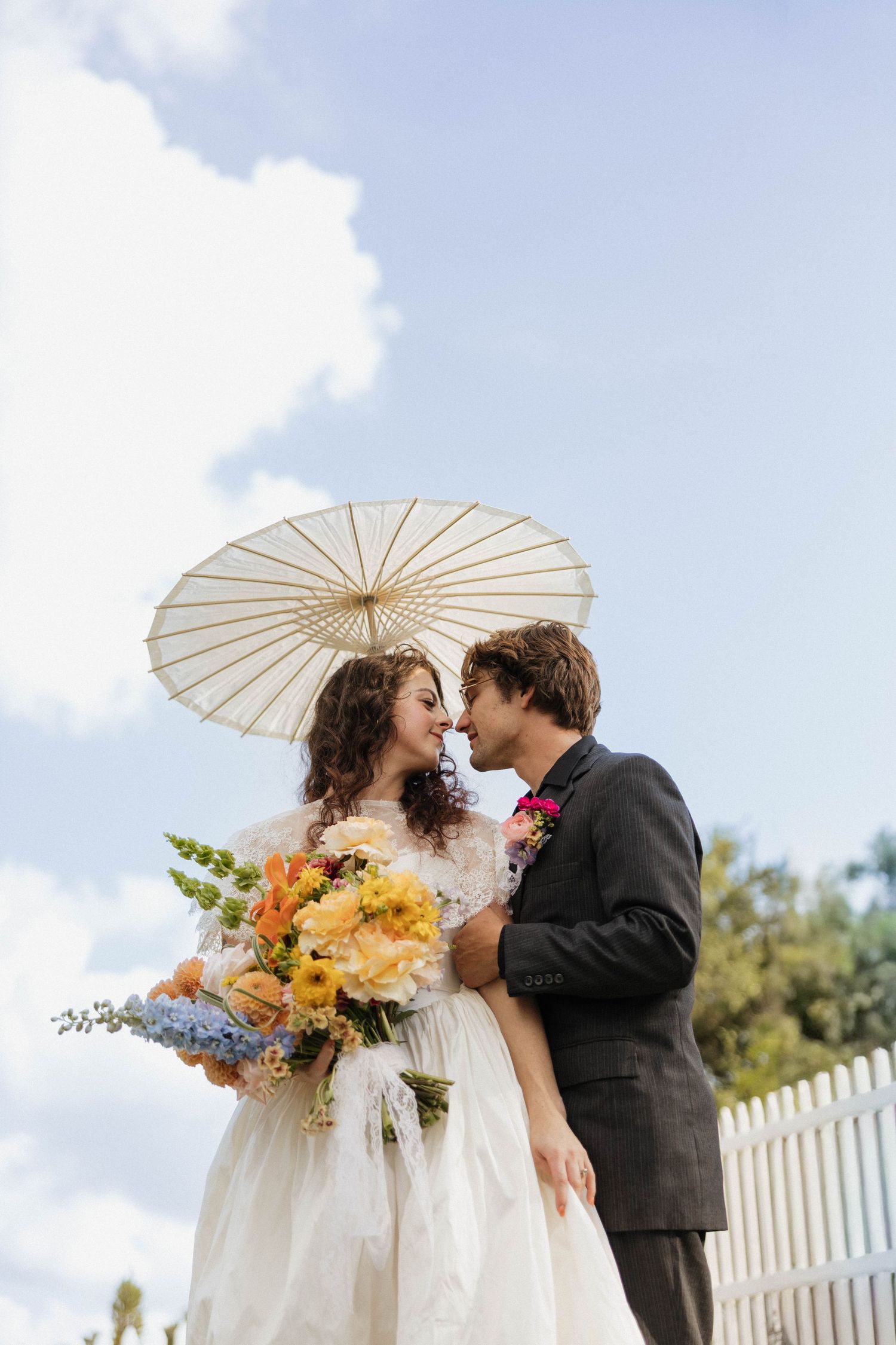 Bride and groom share a romantic moment under a white parasol, bride holding colorful bouquet outdoors.