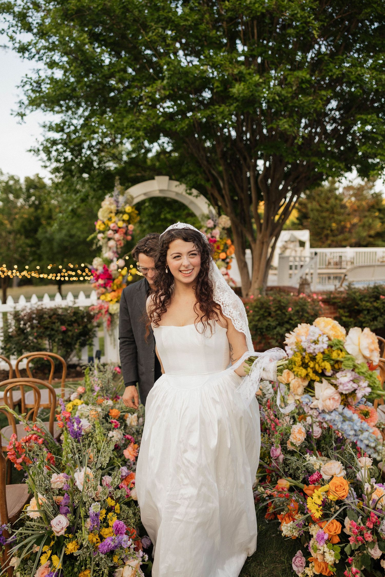 Bride in white strapless gown smiles walking past colorful floral arrangements at outdoor garden wedding ceremony.