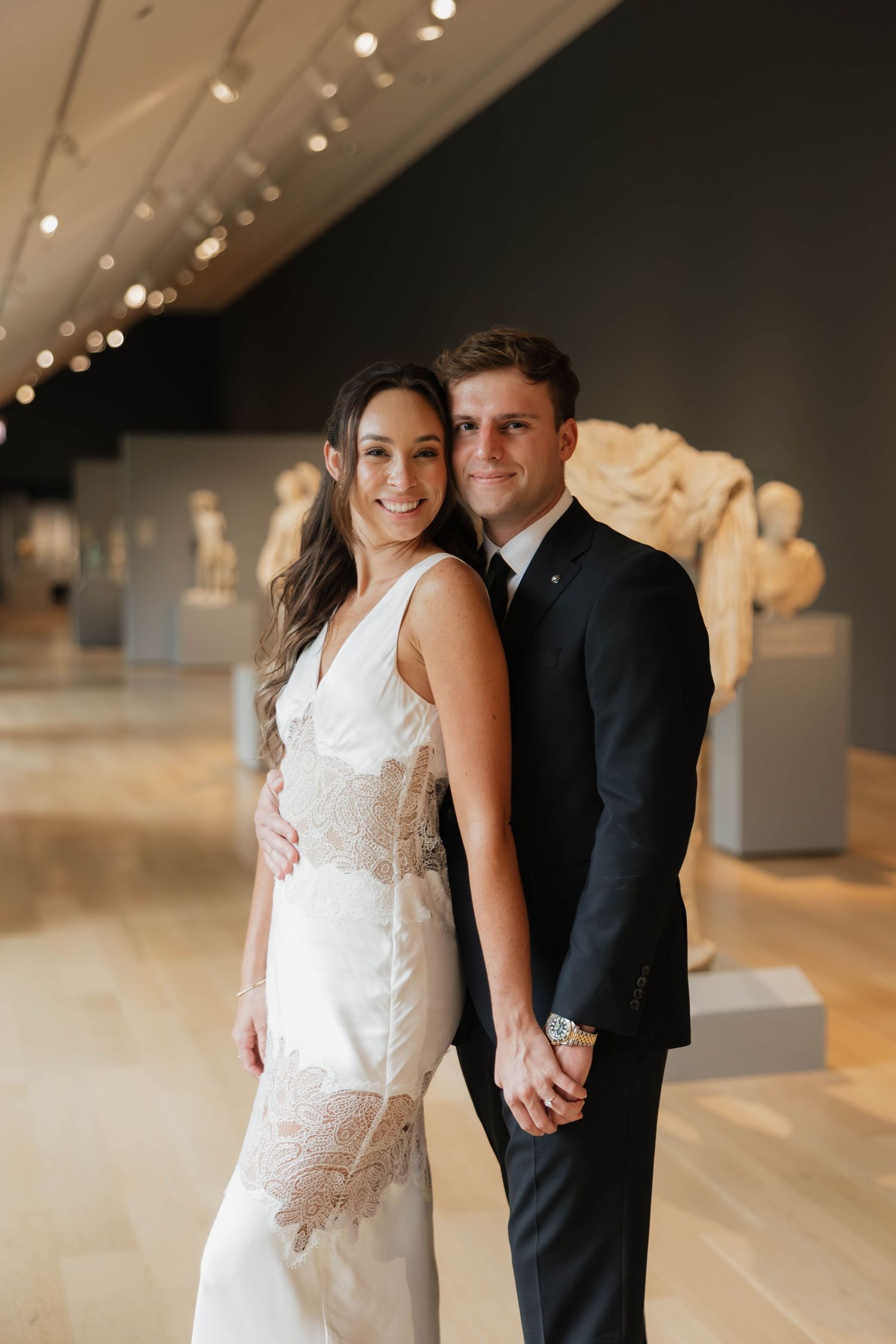 Smiling couple in wedding attire posing in an art museum gallery with classical sculptures in the background.