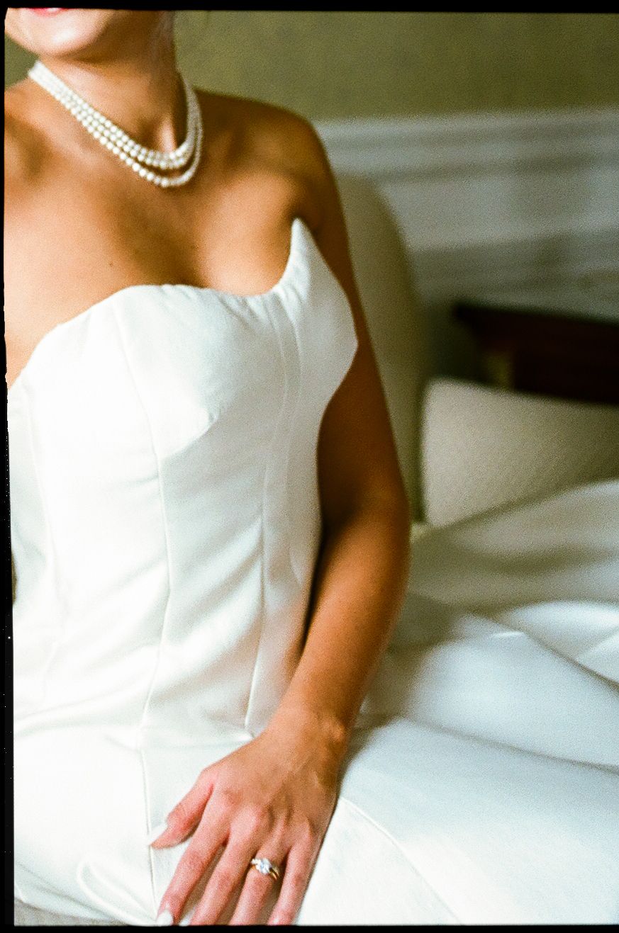 Bride in strapless white gown wearing layered pearl necklace and diamond ring seated on white bedding.