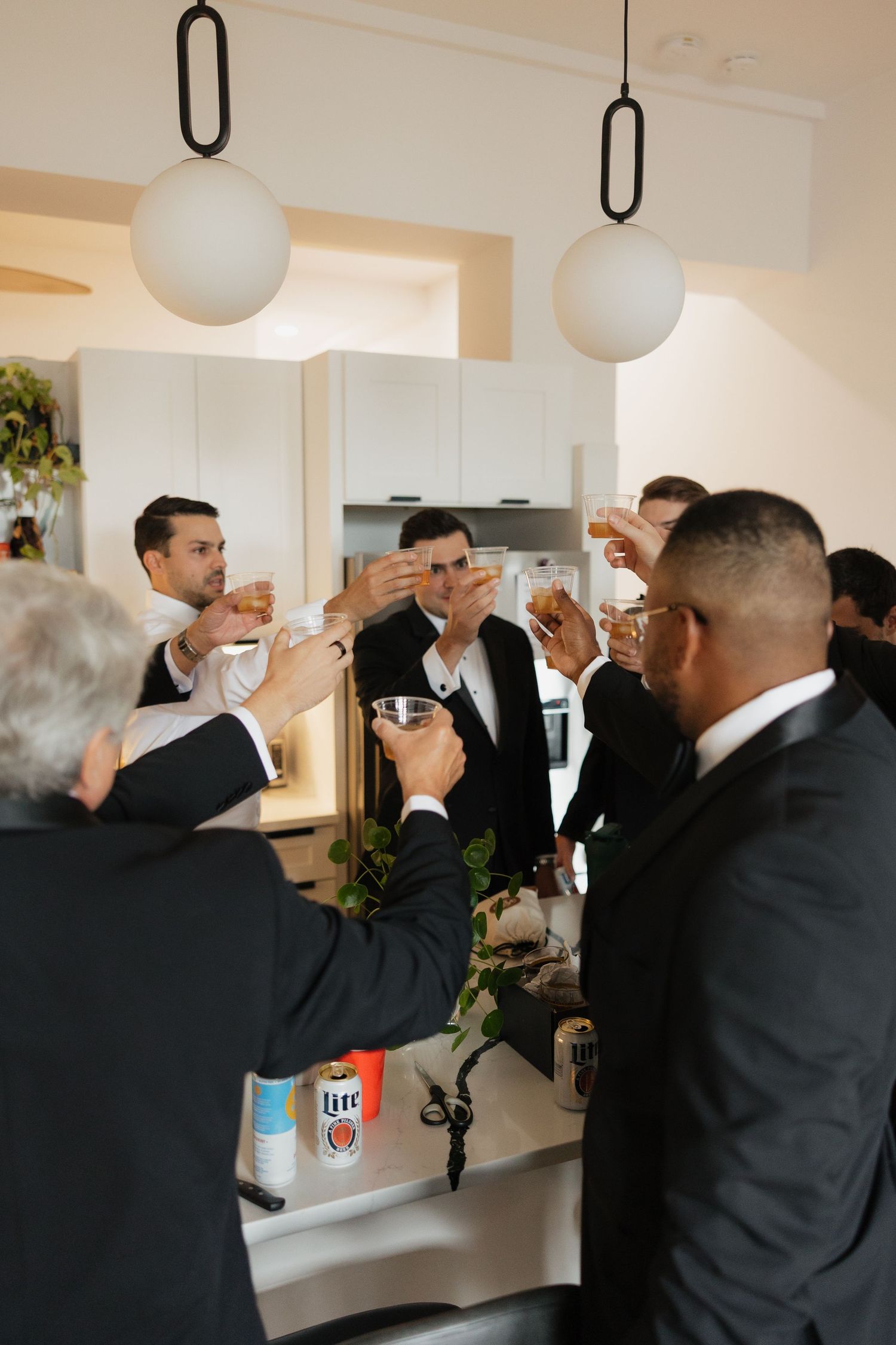 Groomsmen in black suits toasting with drinks in a modern kitchen during a wedding celebration.