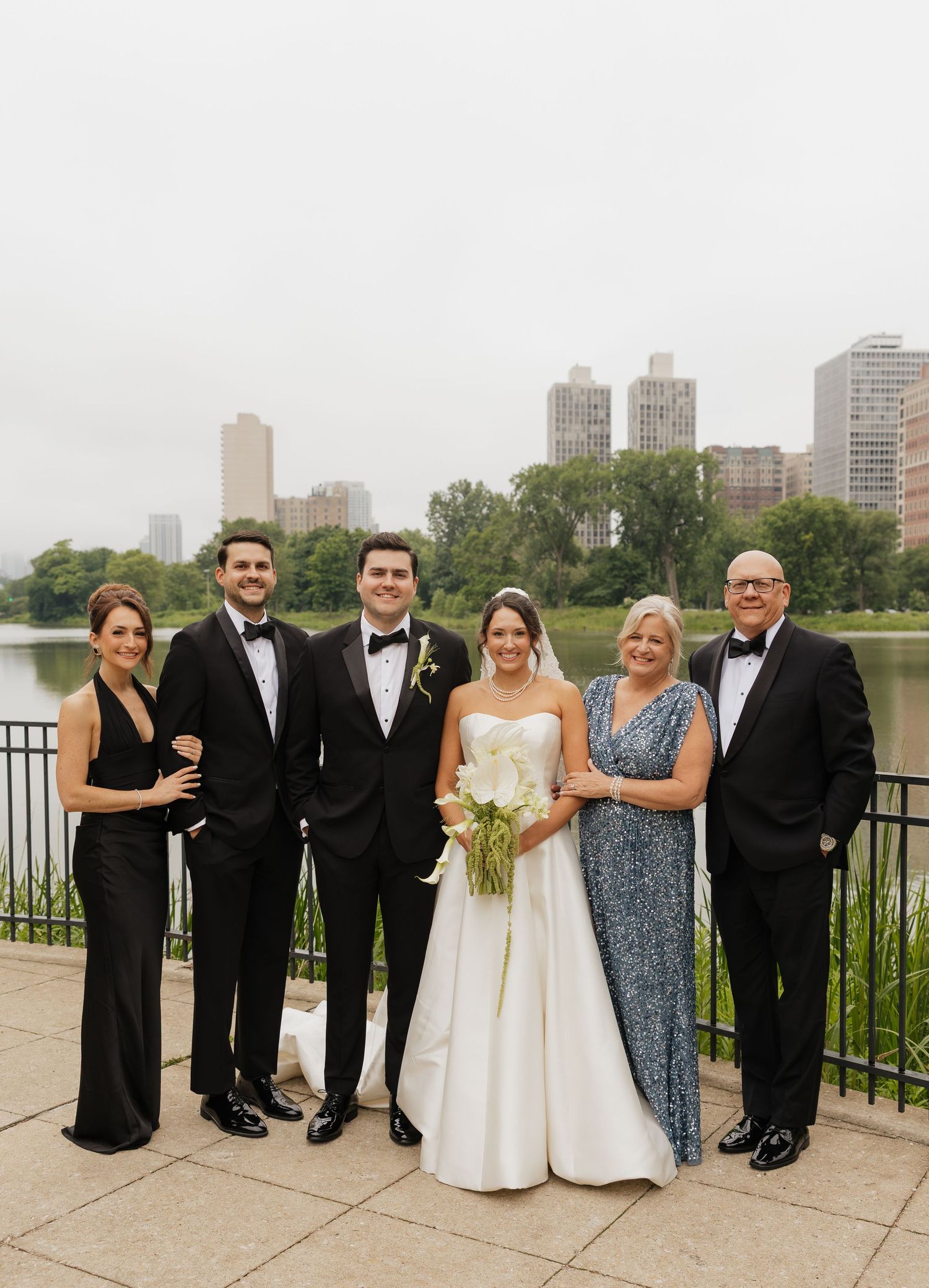 Wedding party of six posing by a lakeside park with city skyline backdrop, bride holding white calla lily bouquet.