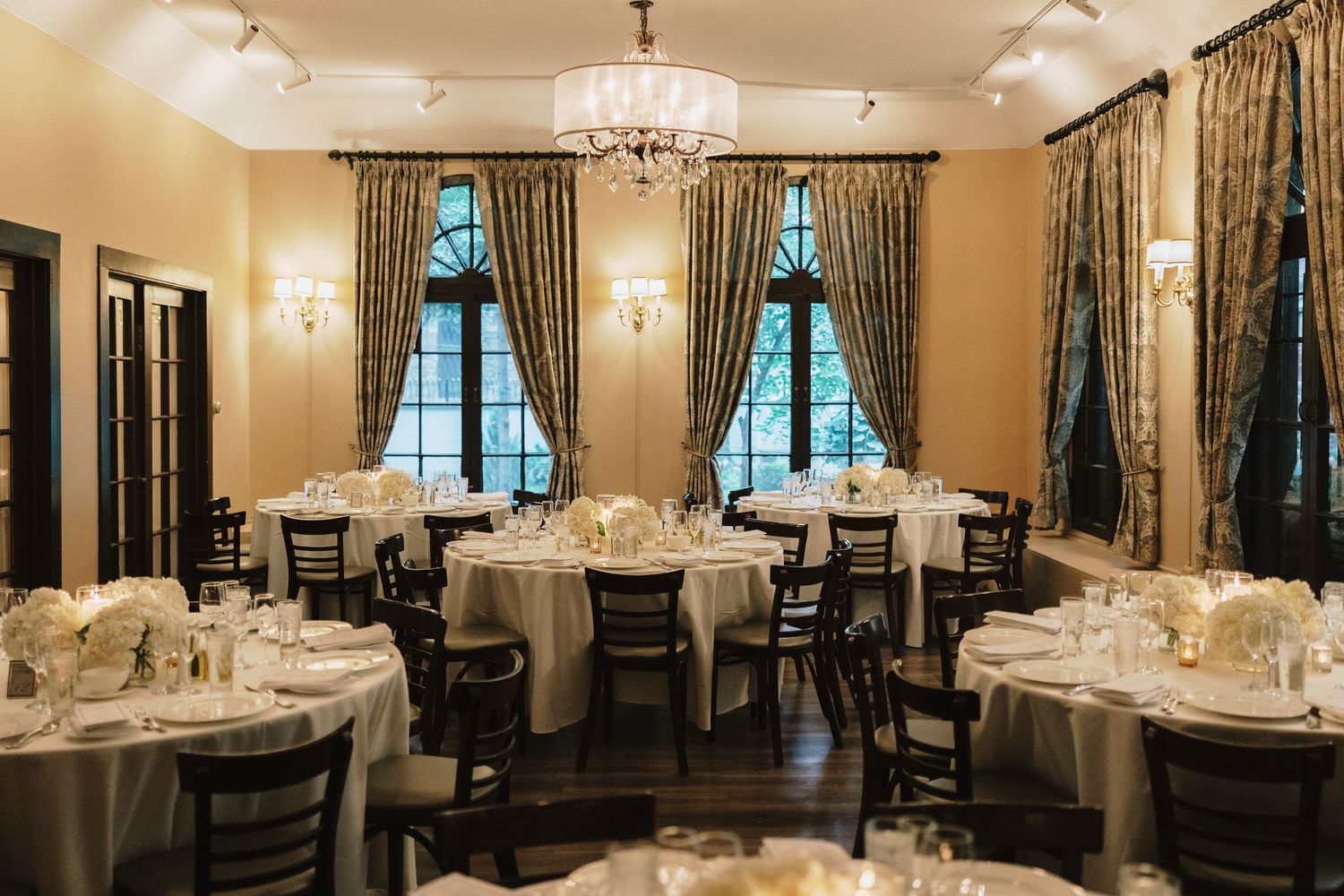 Elegant wedding reception room with round tables, white linens, chandelier, and ornate curtained windows.