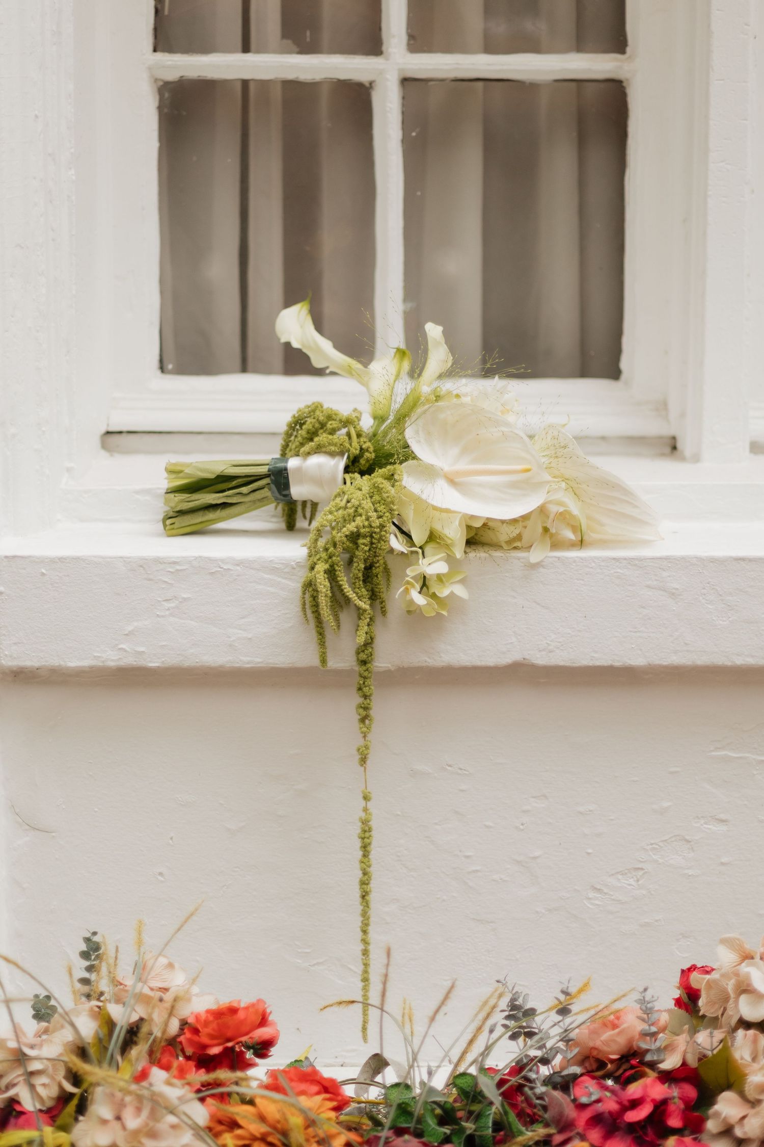 White calla lily bridal bouquet with green amaranthus resting on a white window ledge with colorful flowers below.