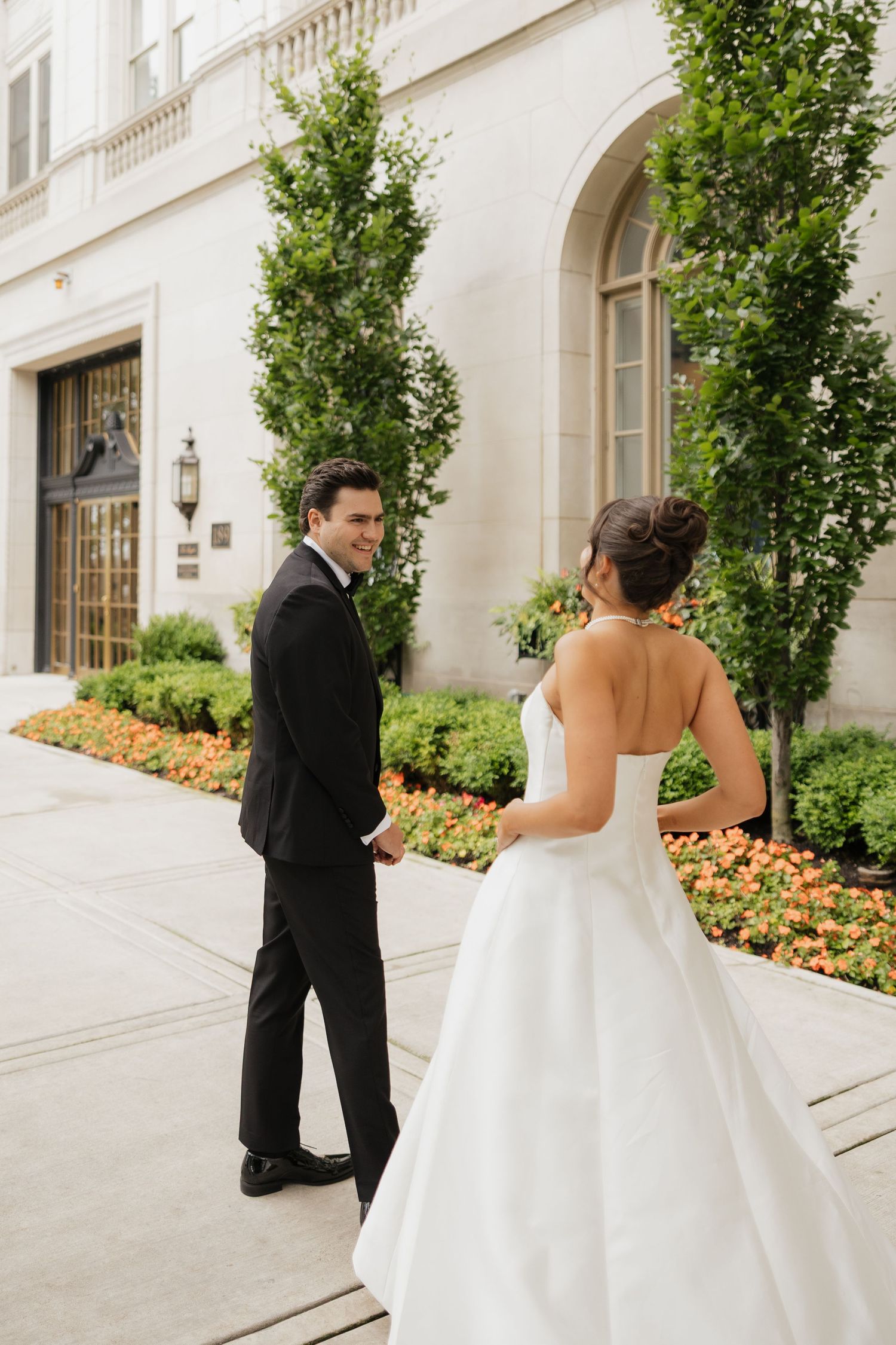 A groom in a black tuxedo smiles back at his bride in a white ball gown outside an elegant building.