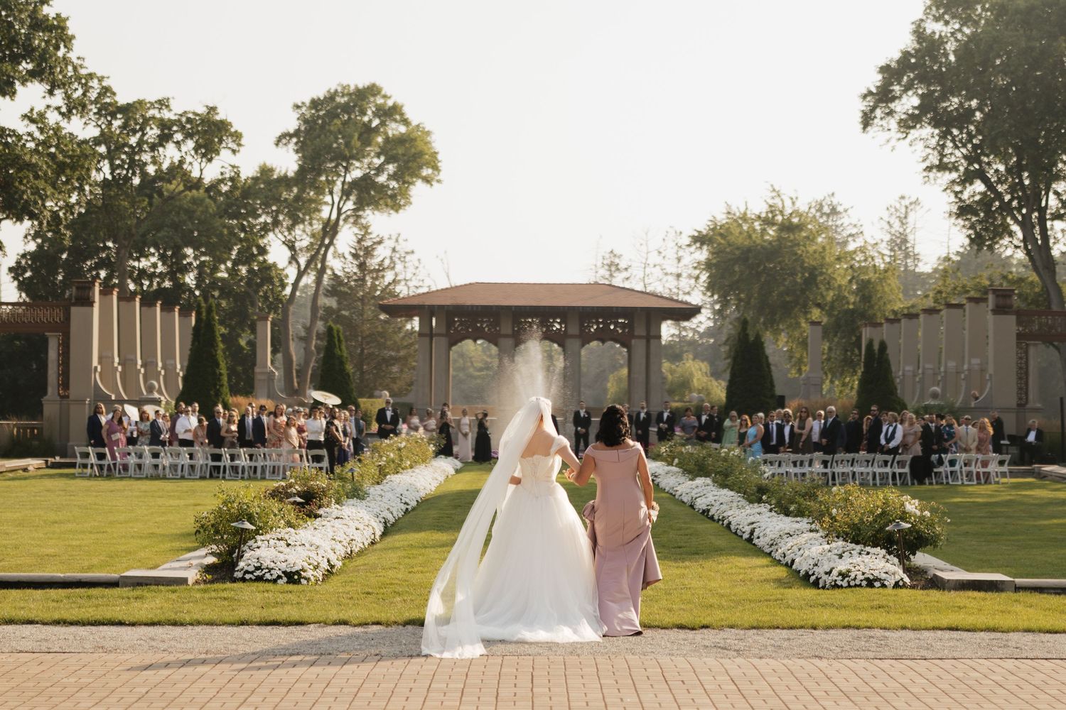 Bride in white gown with veil walks toward outdoor wedding ceremony pavilion surrounded by manicured gardens and seated guests.