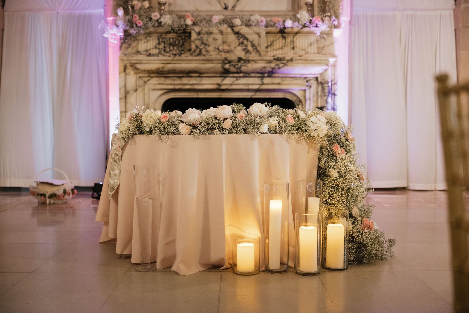 Elegant wedding sweetheart table with white floral garland, candles, and blush draping before ornate fireplace.