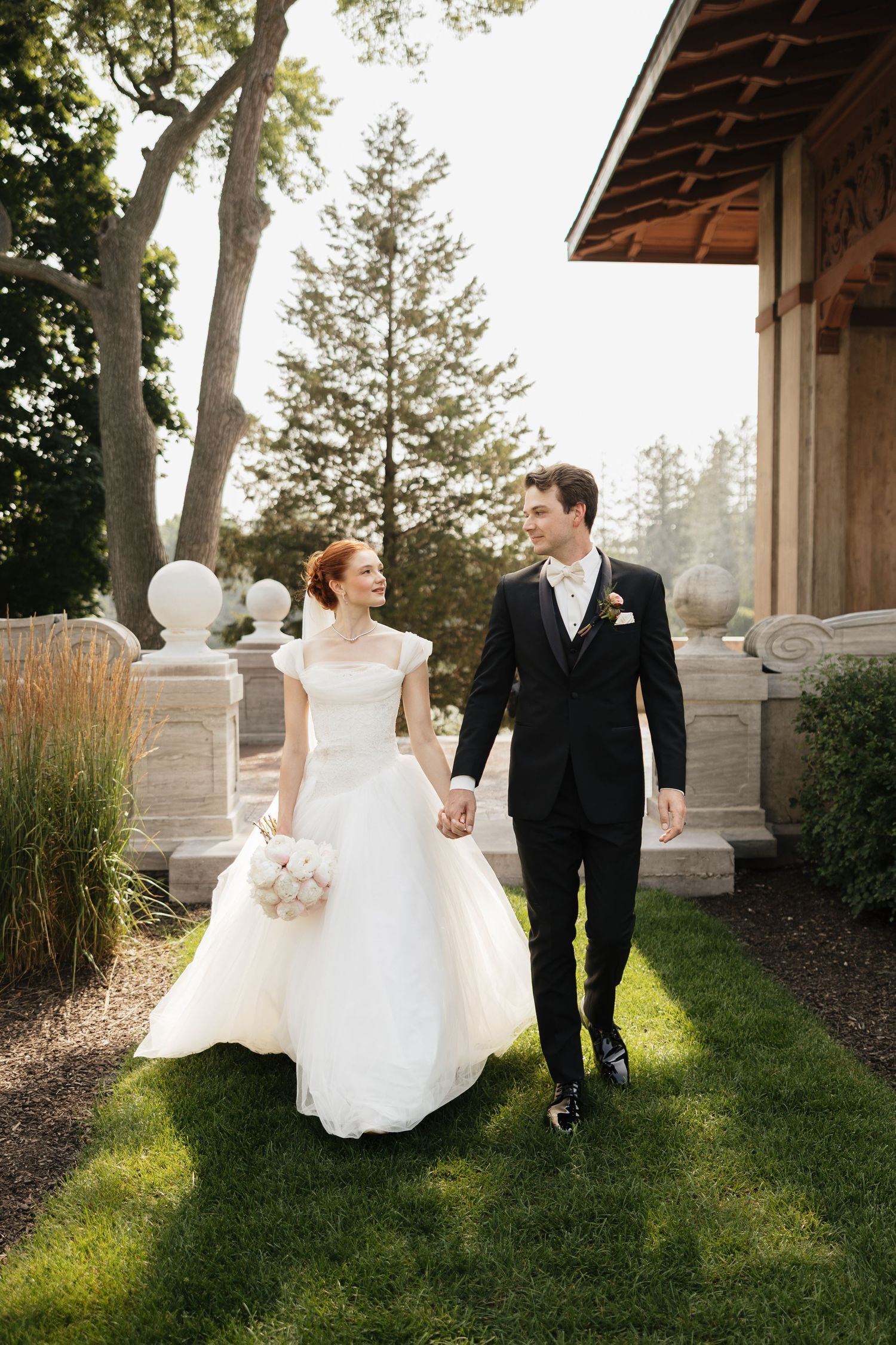 Bride in white gown and groom in black tuxedo walk hand-in-hand on green lawn outside elegant venue.