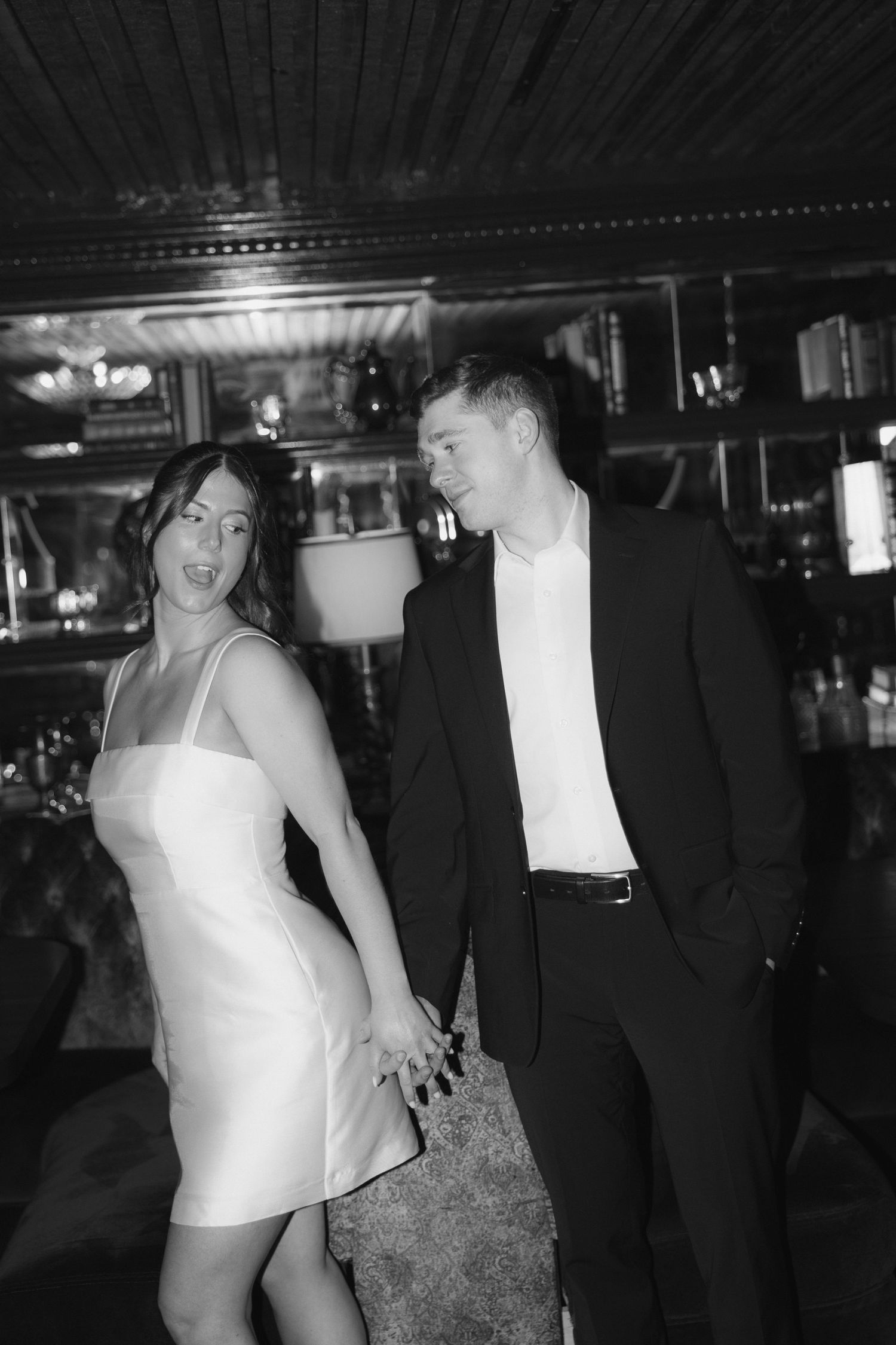 A couple in formal attire, with a white dress and black suit, share an intimate moment in a dimly lit bar setting.