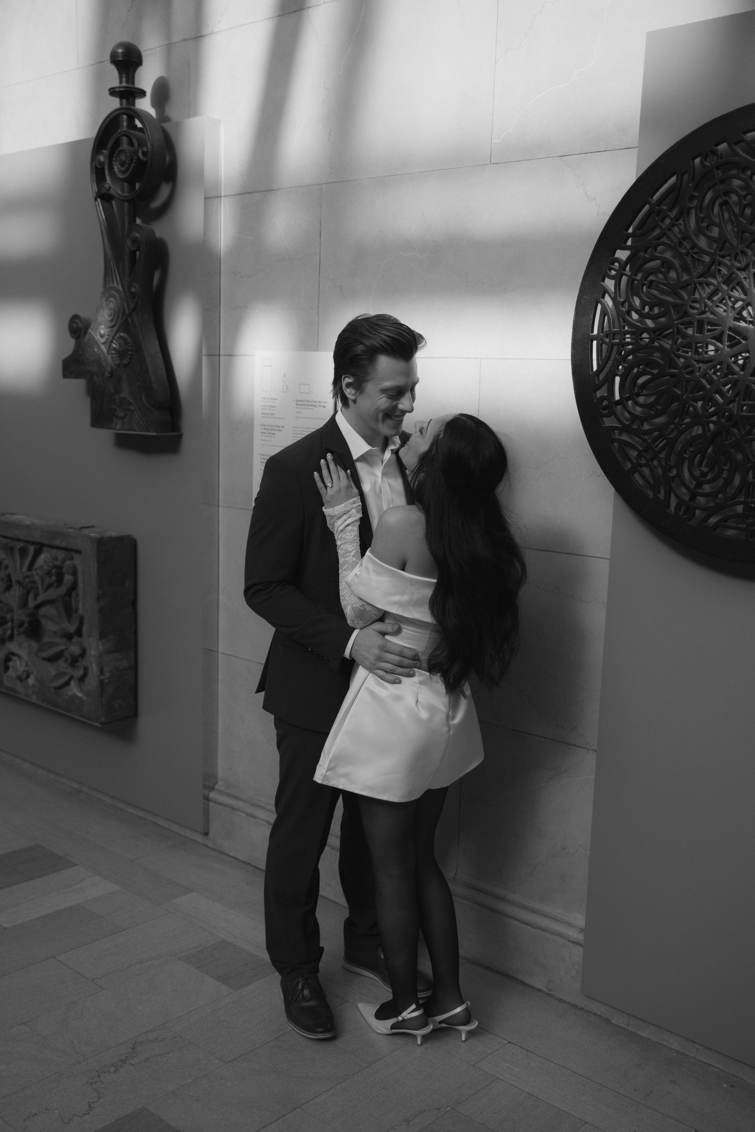 Two people share an intimate black and white moment against a wall with decorative art pieces.
