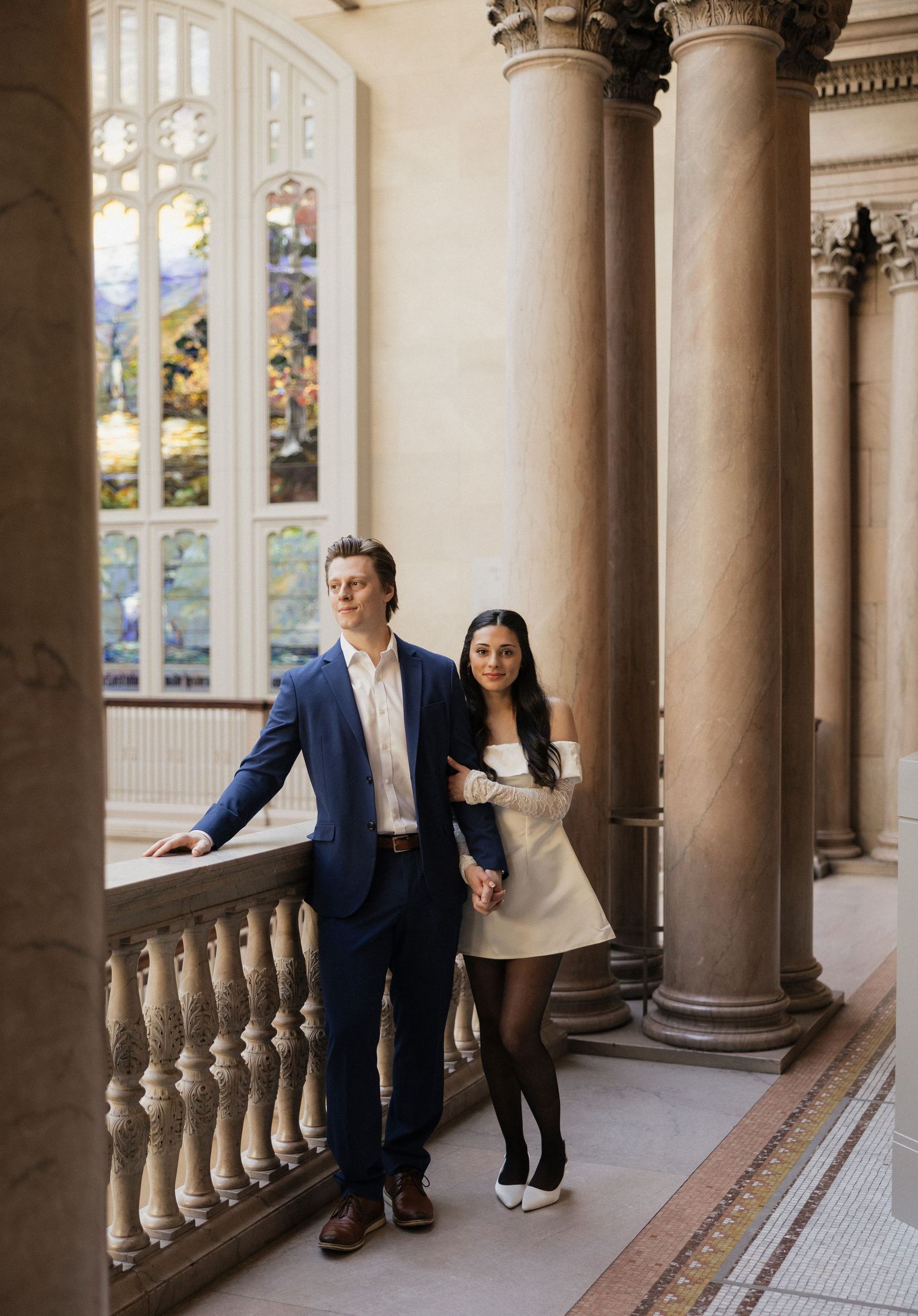 A couple poses in an elegant hallway with classical architecture and stained glass windows.